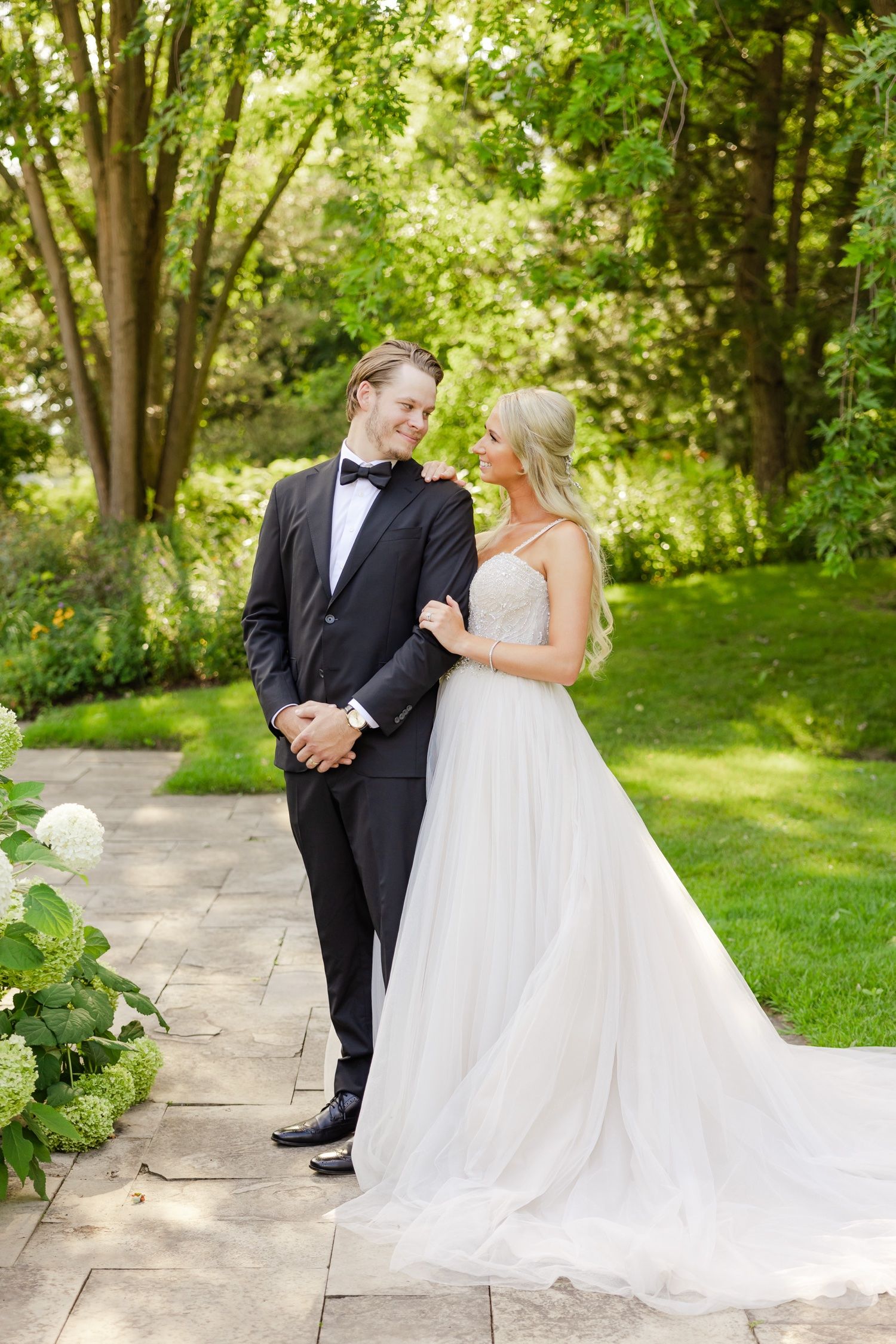Elegant wedding portrait on stone pathway with white hydrangeas and green trees.