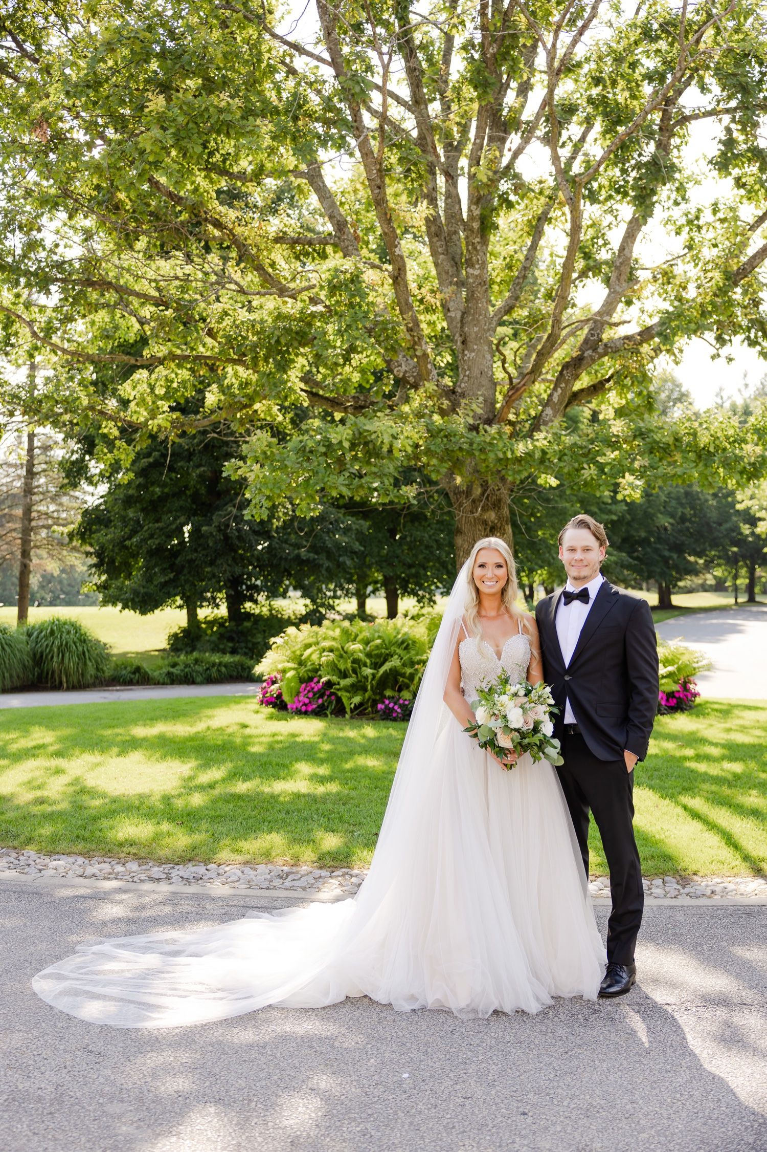 Bride and groom pose for a formal wedding portrait in a sunlit garden setting.