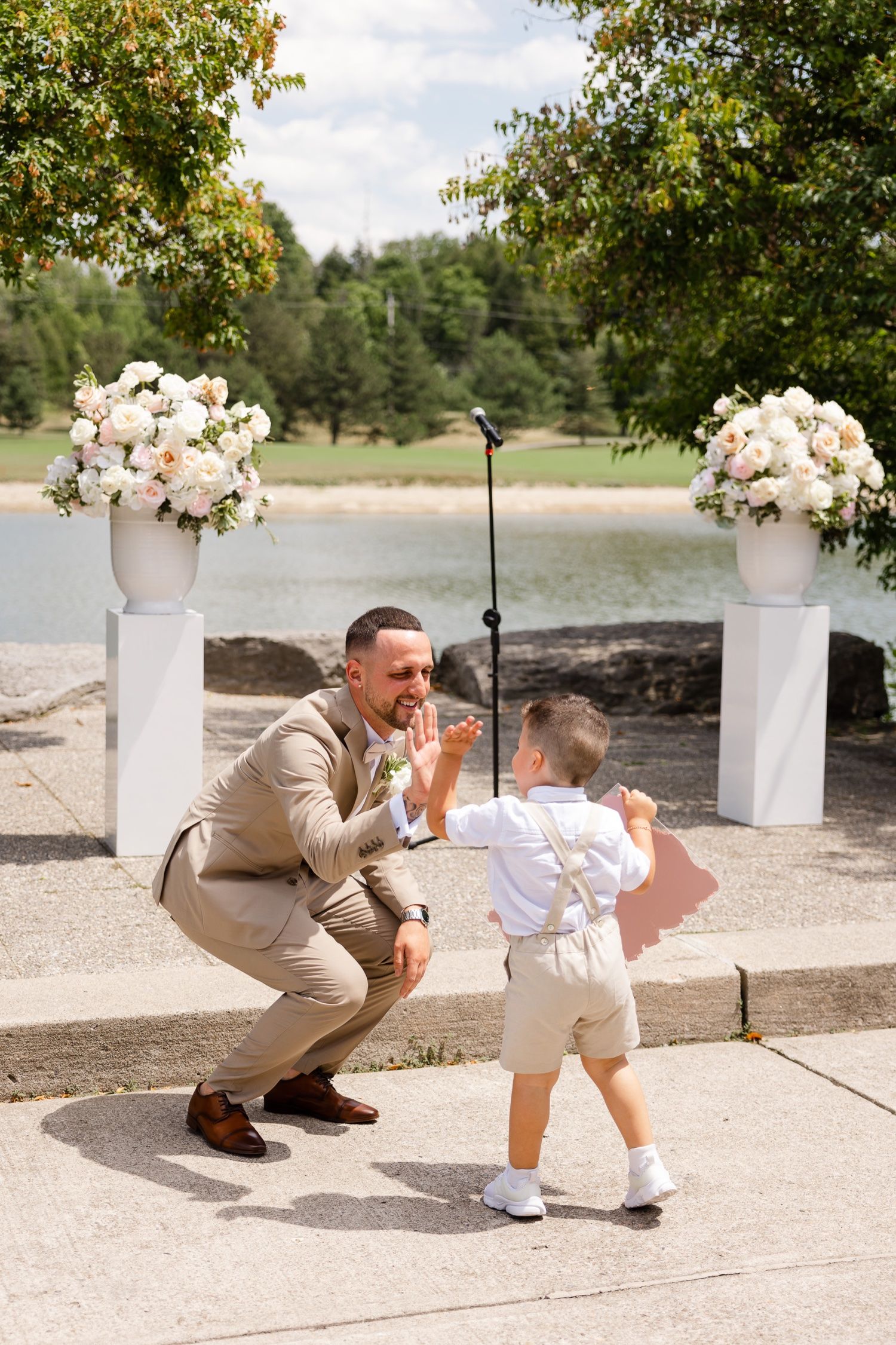 A heartwarming moment between groom in beige suit and young ring bearer at lakeside wedding ceremony.
