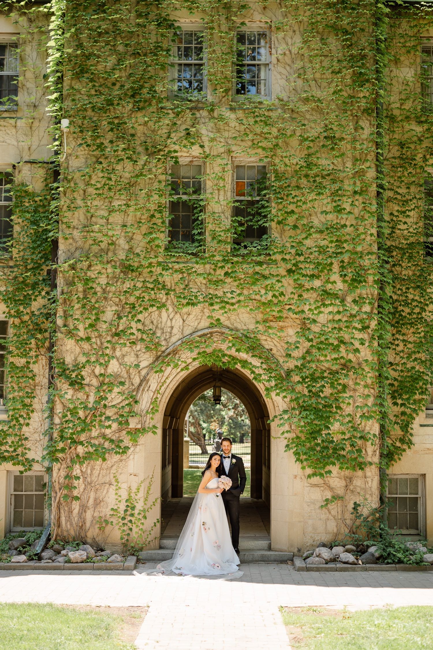 Wedding portrait under ivy-covered stone archway of historic university building.