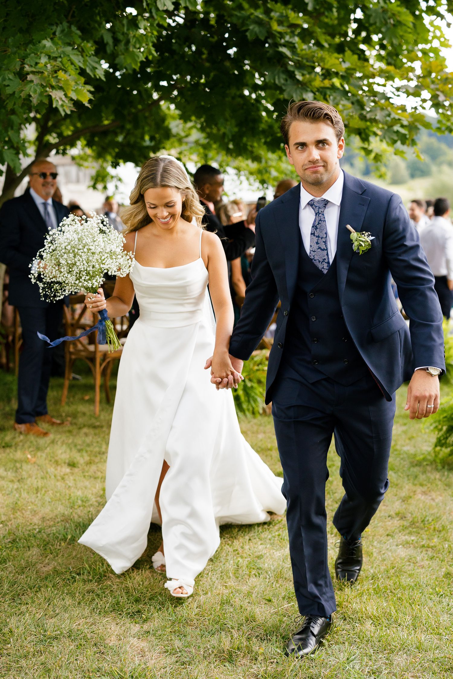 Newlyweds walking together on grass under trees at an outdoor wedding celebration.