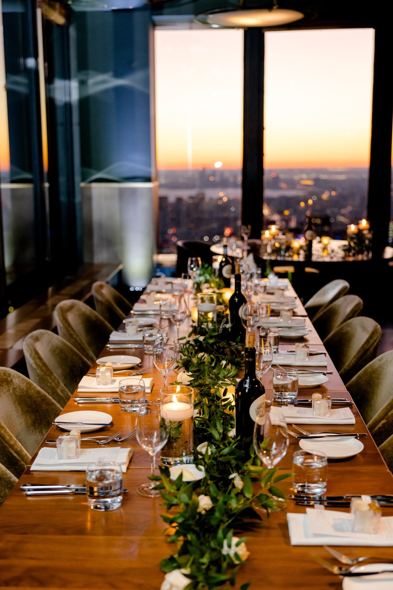 Long dining table set for wedding reception with city sunset views through floor-to-ceiling windows.