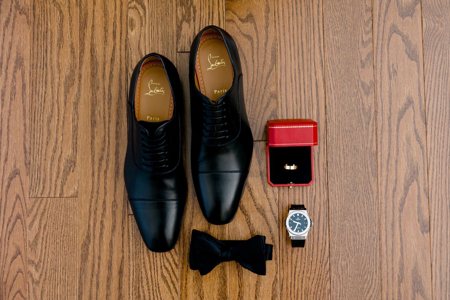 Black dress shoes with bow tie, ring box and watch laid out on wooden floor.