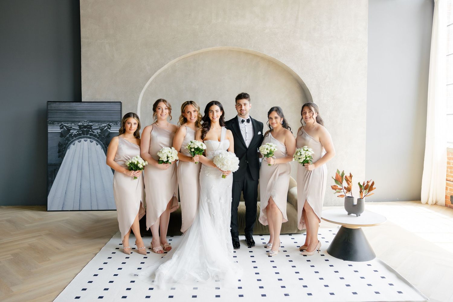 Wedding party in blush dresses poses with couple under arched alcove.