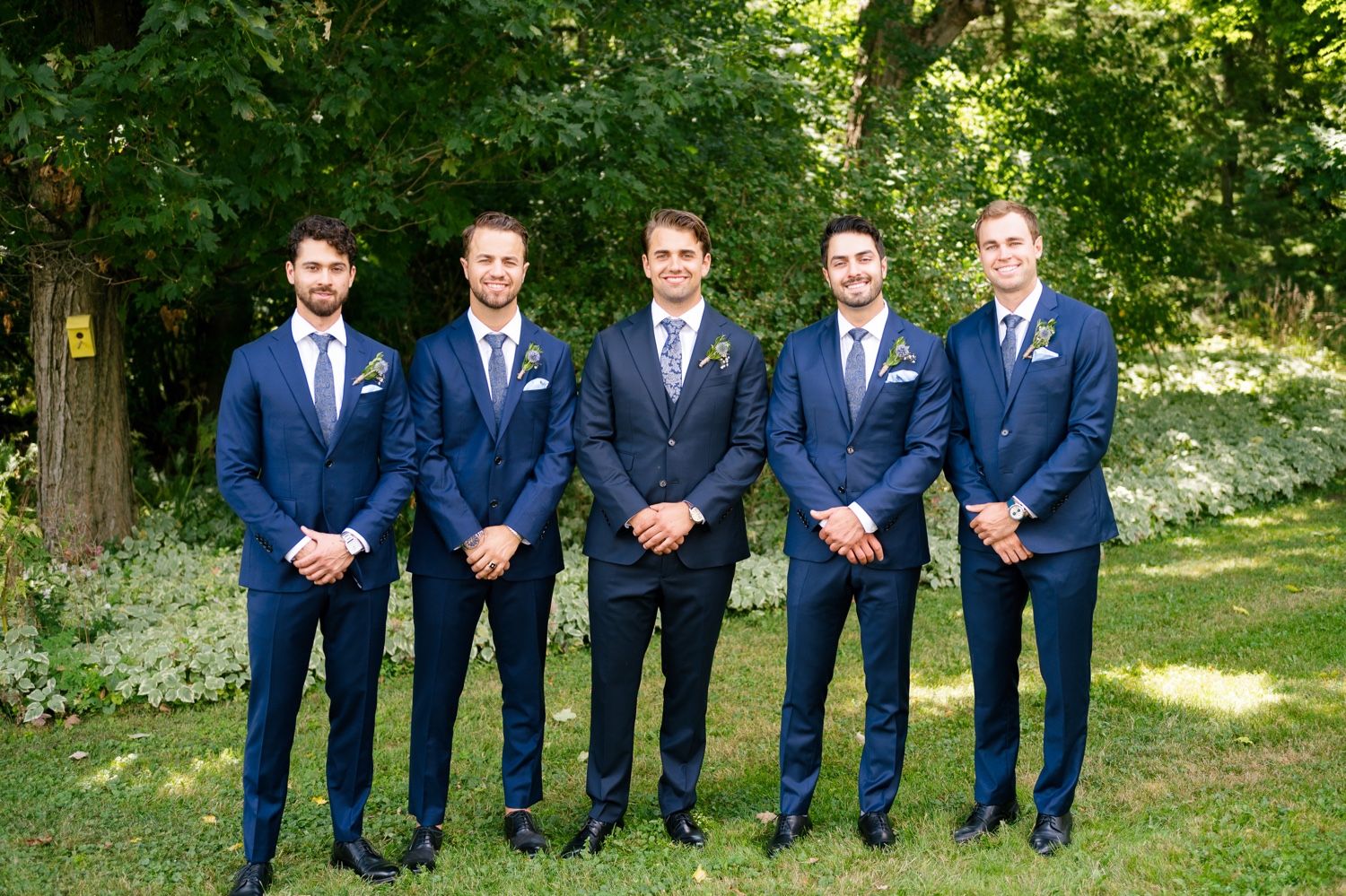 Group of groomsmen in coordinated navy suits lined up for formal photo in garden setting.