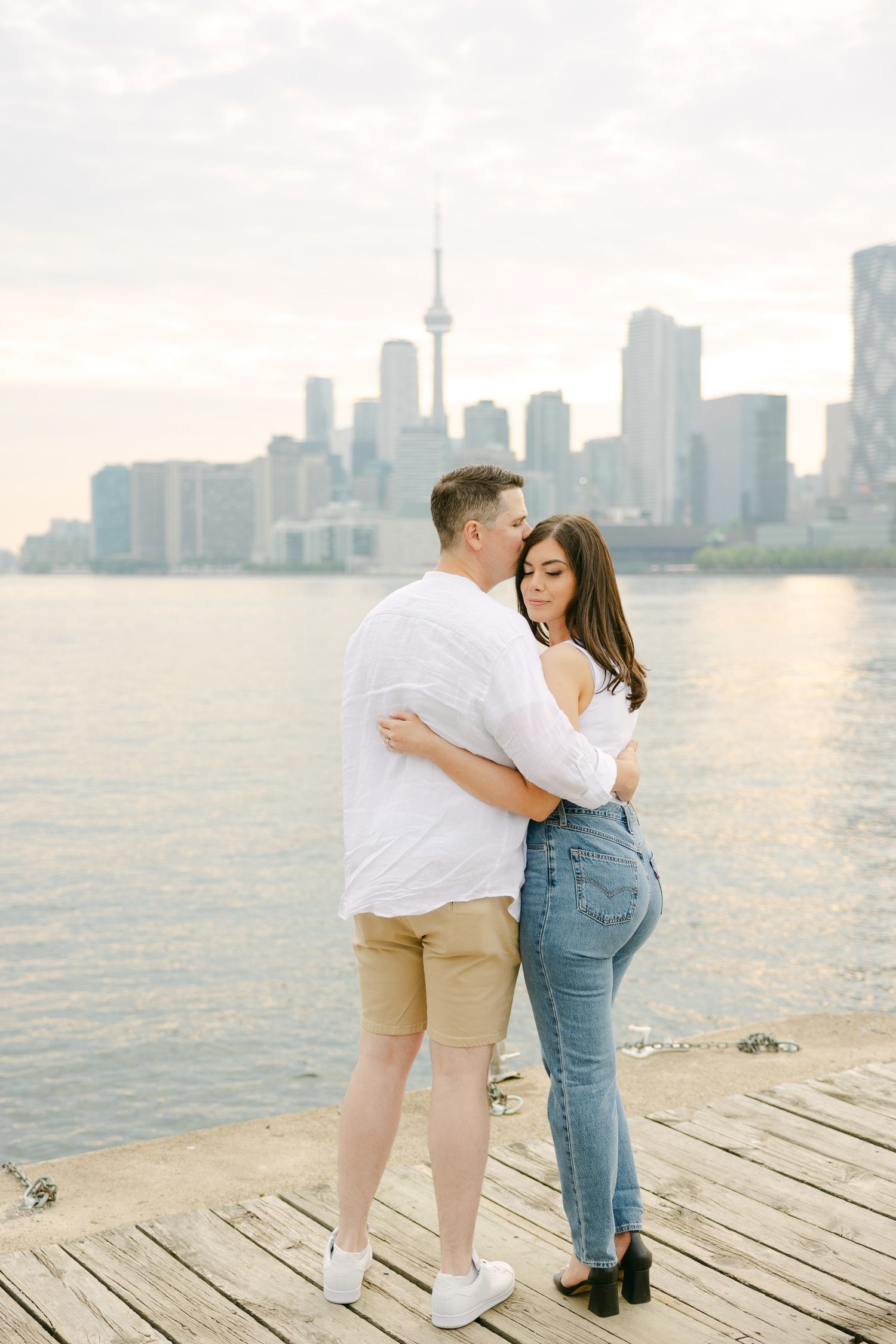 A couple stands close on a wooden dock with Toronto's waterfront skyline in soft evening light.
