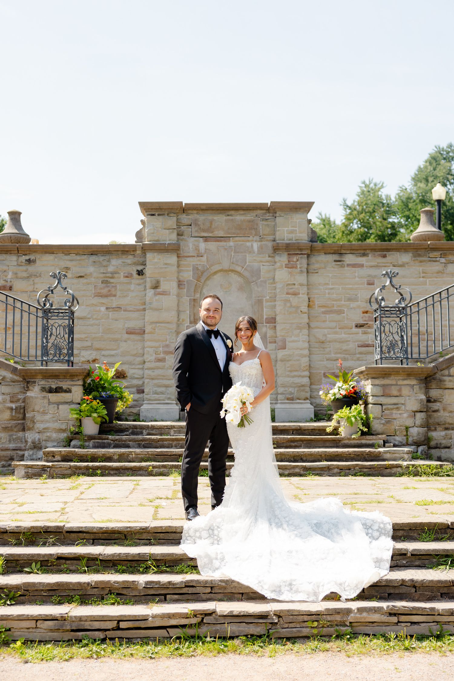 A newlywed couple stands on grand stone steps in front of an ornate architectural entrance with beautiful flower arrangements.
