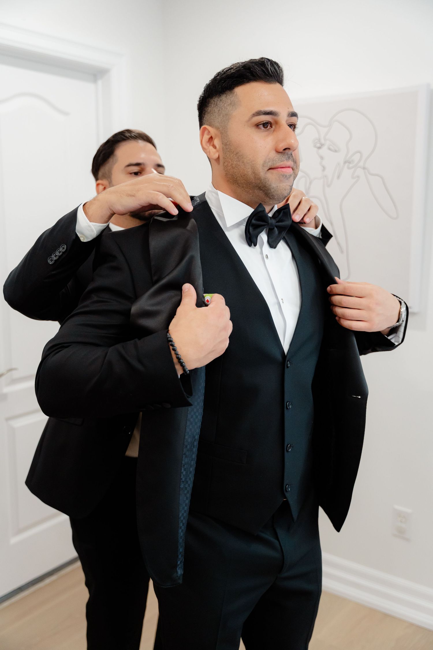A groomsman helps adjust the suit jacket of the groom during wedding preparations in a bright room.