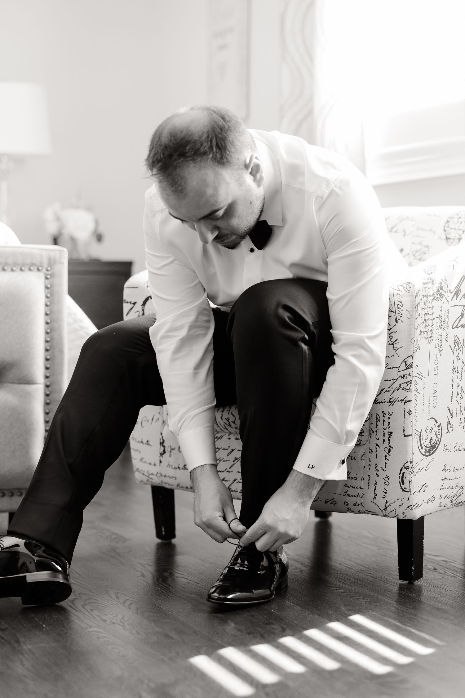 A person in formal attire wearing a white dress shirt and bow tie sits in a chair while tying dress shoes.