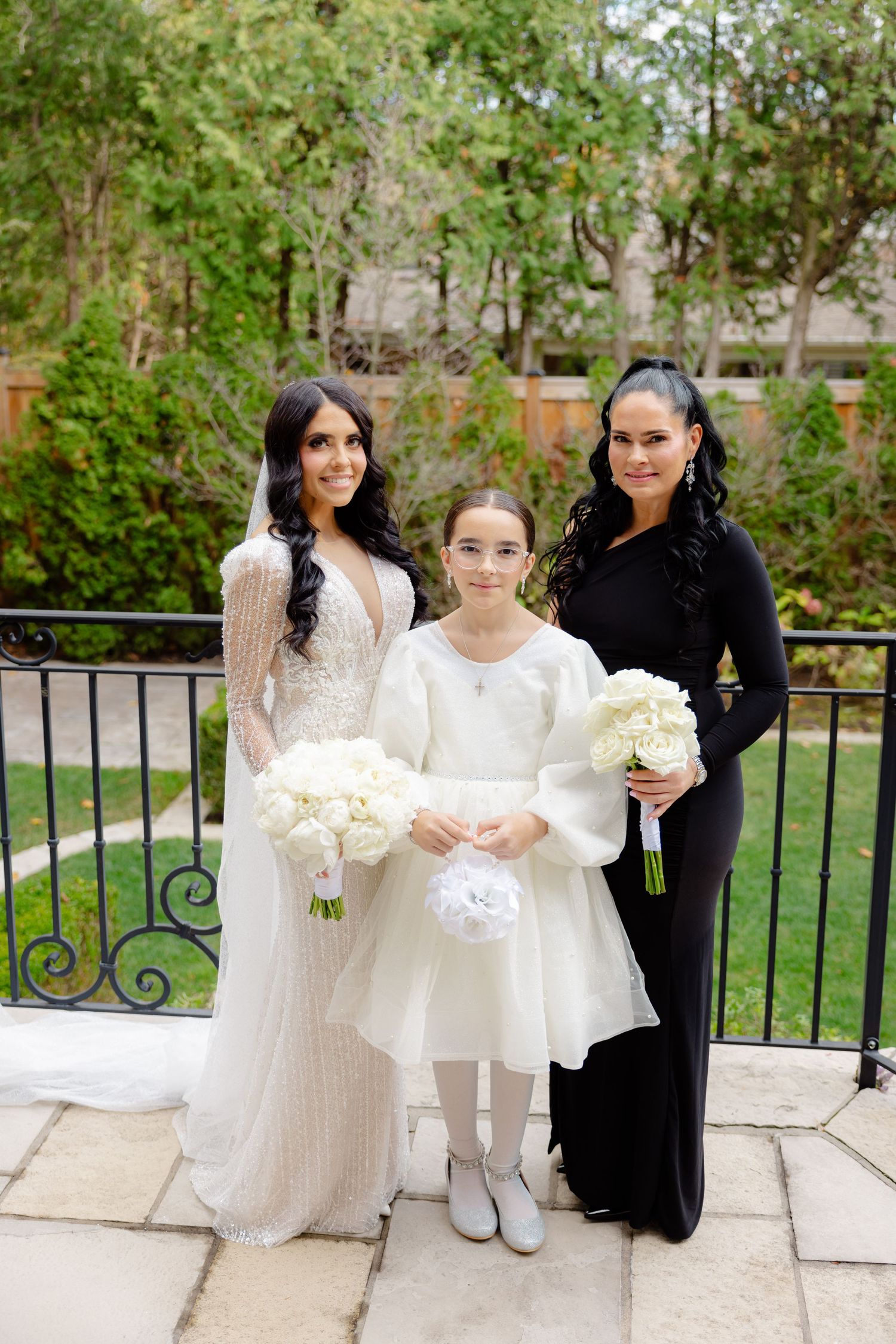 Wedding party poses on stone patio with black iron railing, wearing white and black formal attire.