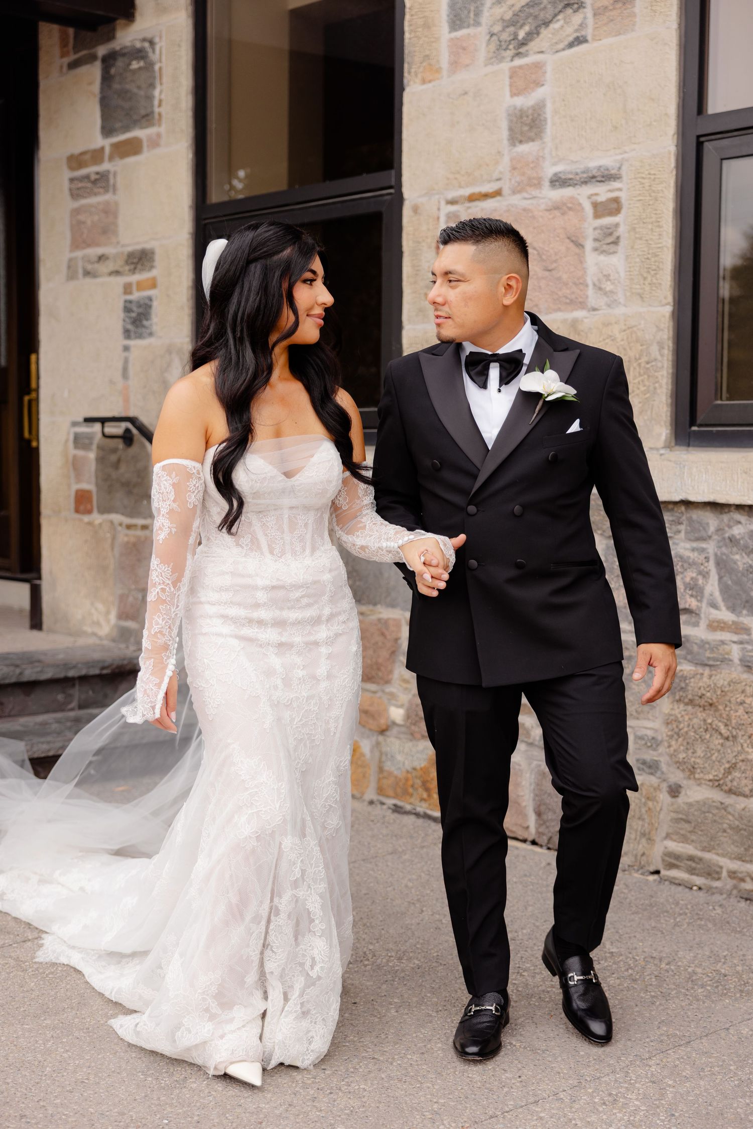 A newlywed couple walks hand in hand along a stone building exterior during their wedding photoshoot.
