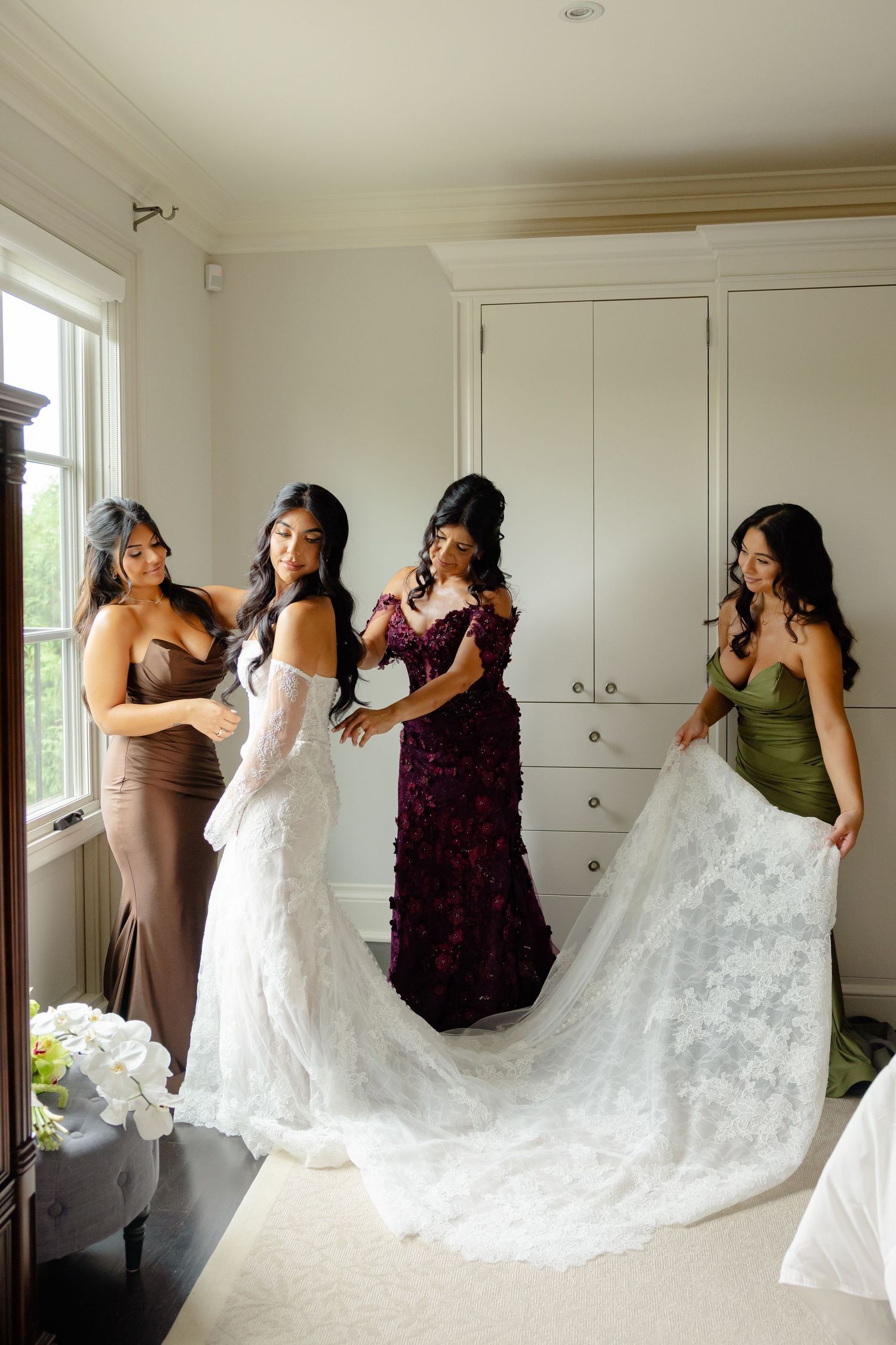 In a white room, five women help adjust the train of an elegant white lace wedding dress during pre-ceremony preparations.
