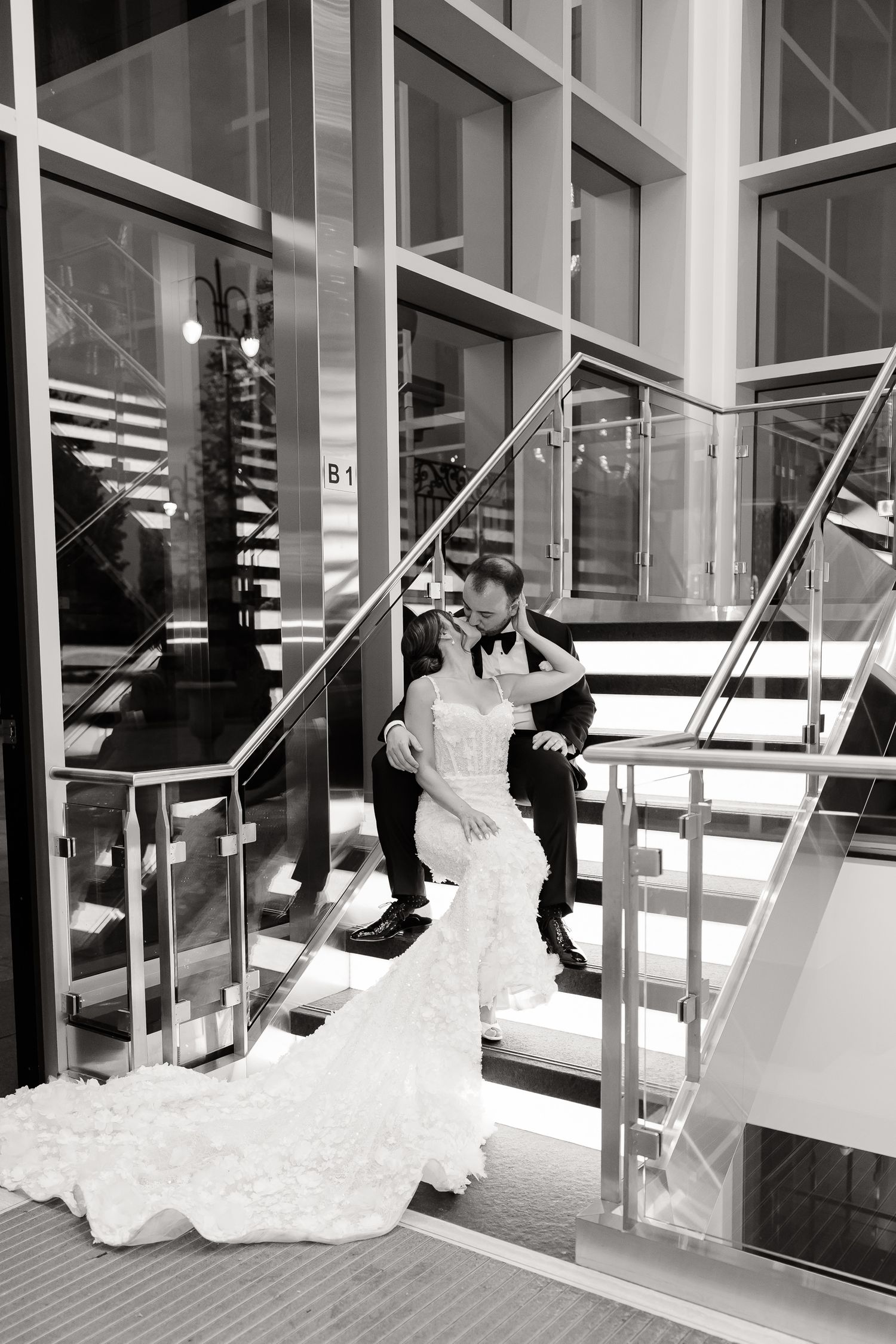A bride in a stunning mermaid gown sits with a companion on modern glass staircase in black and white photography.