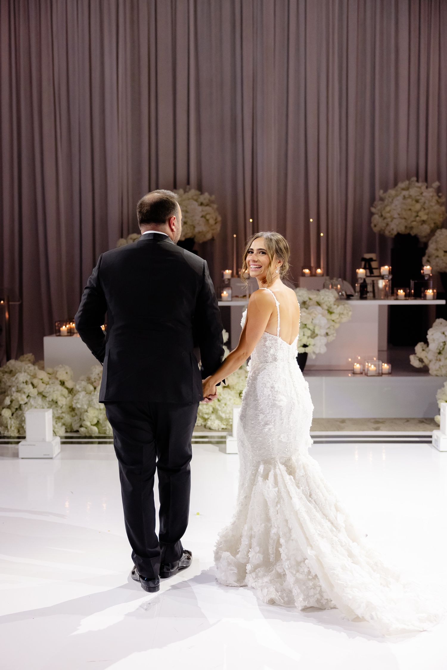A bride in a lace mermaid wedding gown and groom in black tuxedo stand on a white dance floor with floral decor.