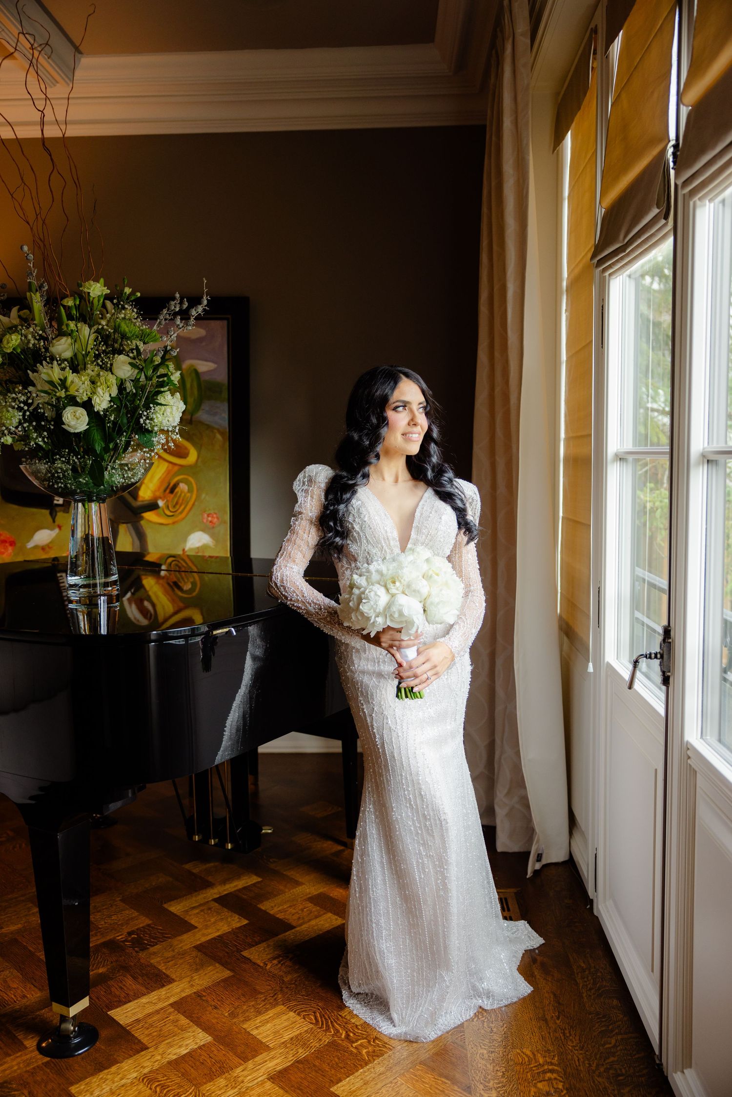 Bride in fitted lace gown holding white bouquet poses by window near grand piano.