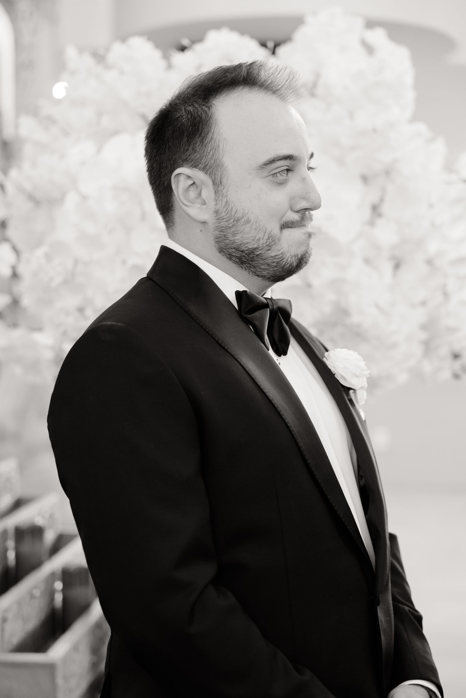 Black and white portrait of groom in tuxedo with white boutonniere at wedding ceremony.