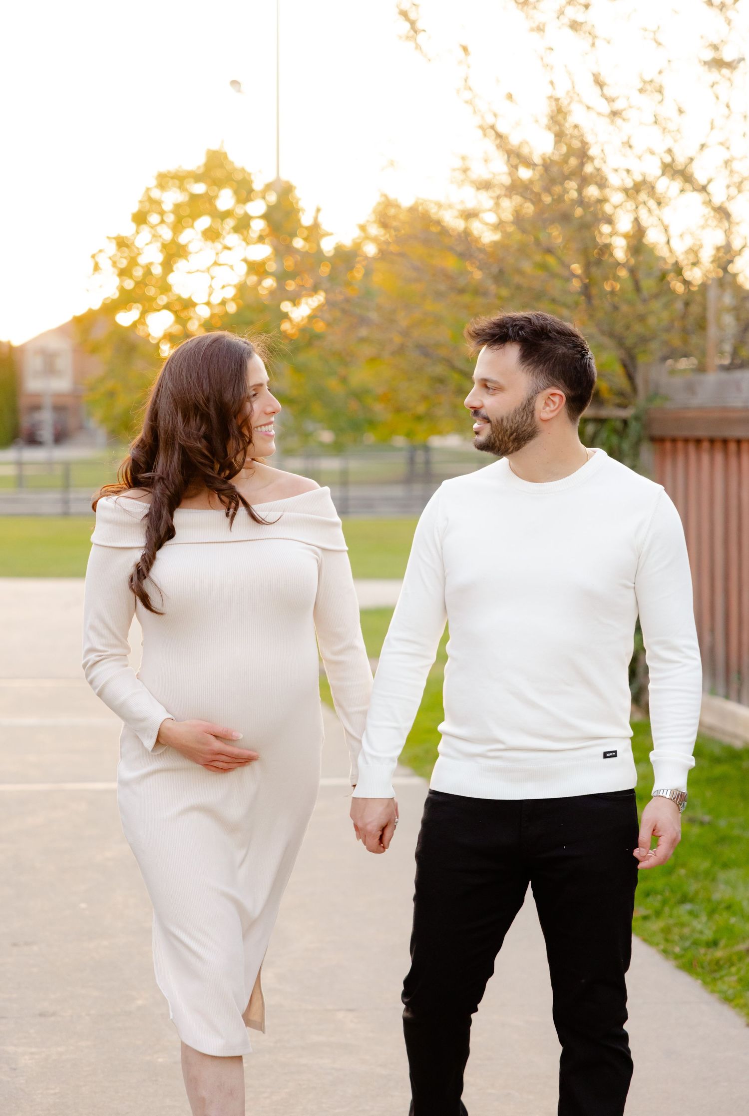 A couple walks hand in hand on a path in a park during golden hour, with the woman wearing a white dress.