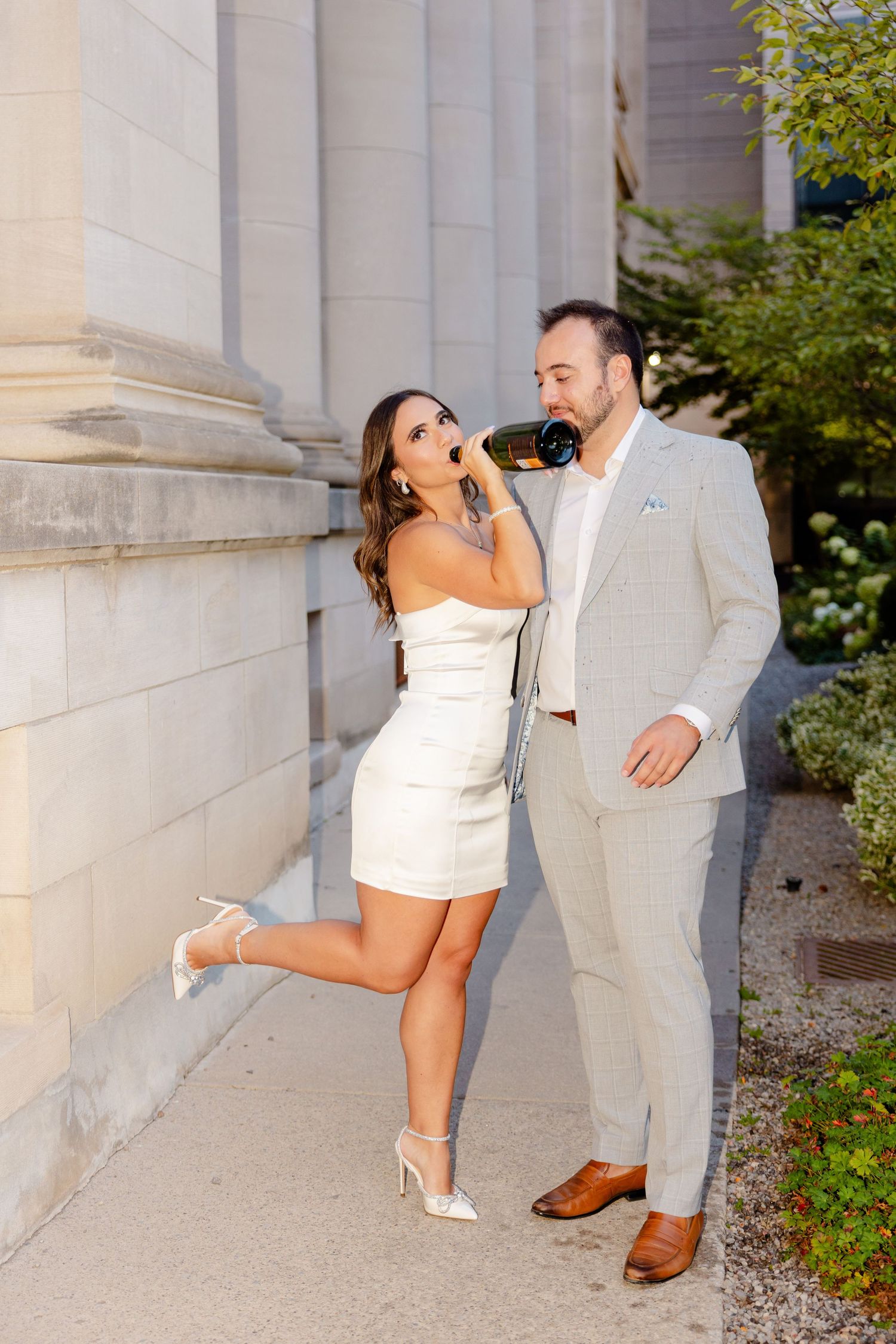 A couple in formal attire poses playfully against a stone column, with her in a white dress and him in a light gray suit.