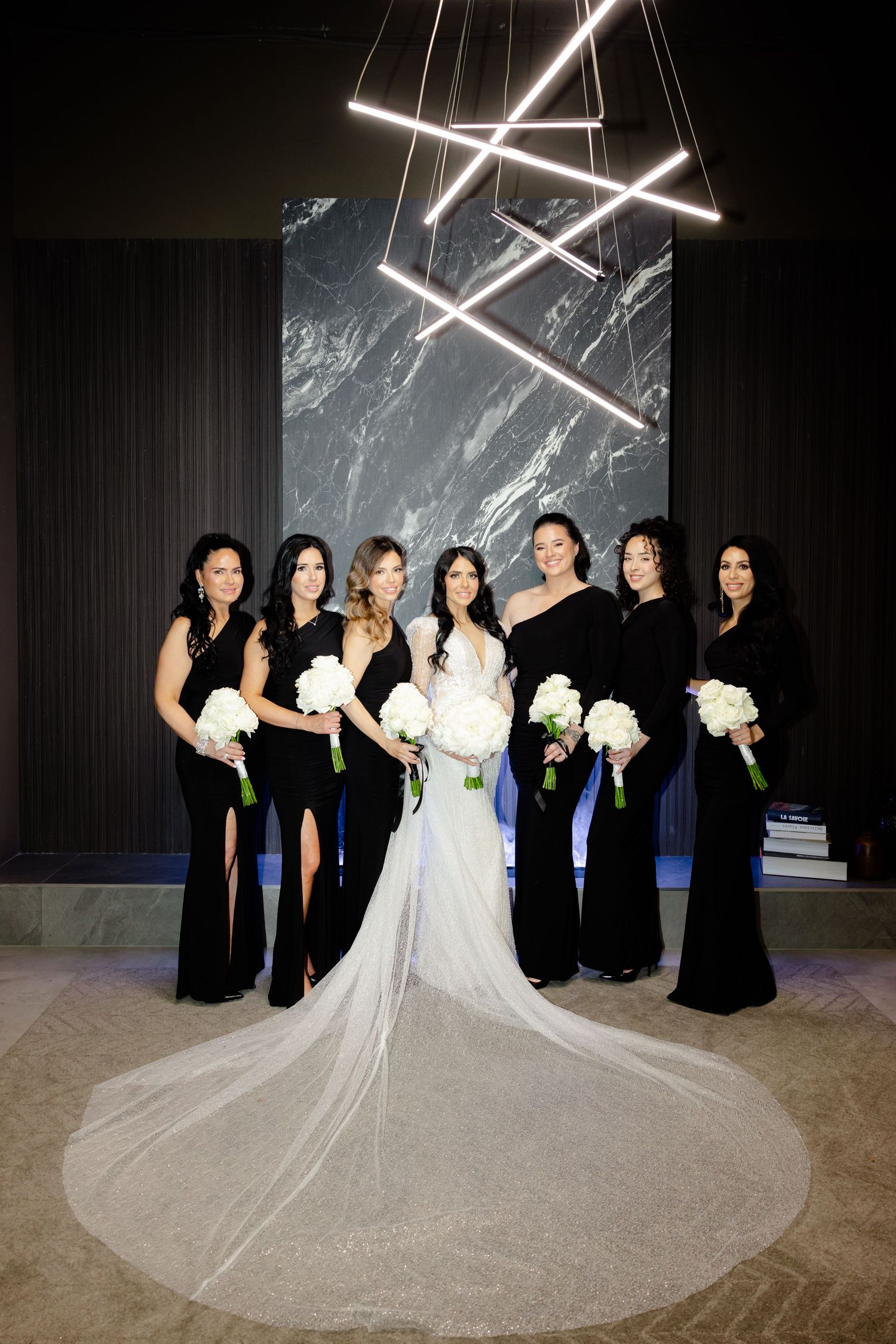 Bridesmaids in black gowns hold white bouquets beside bride with dramatic cathedral veil.