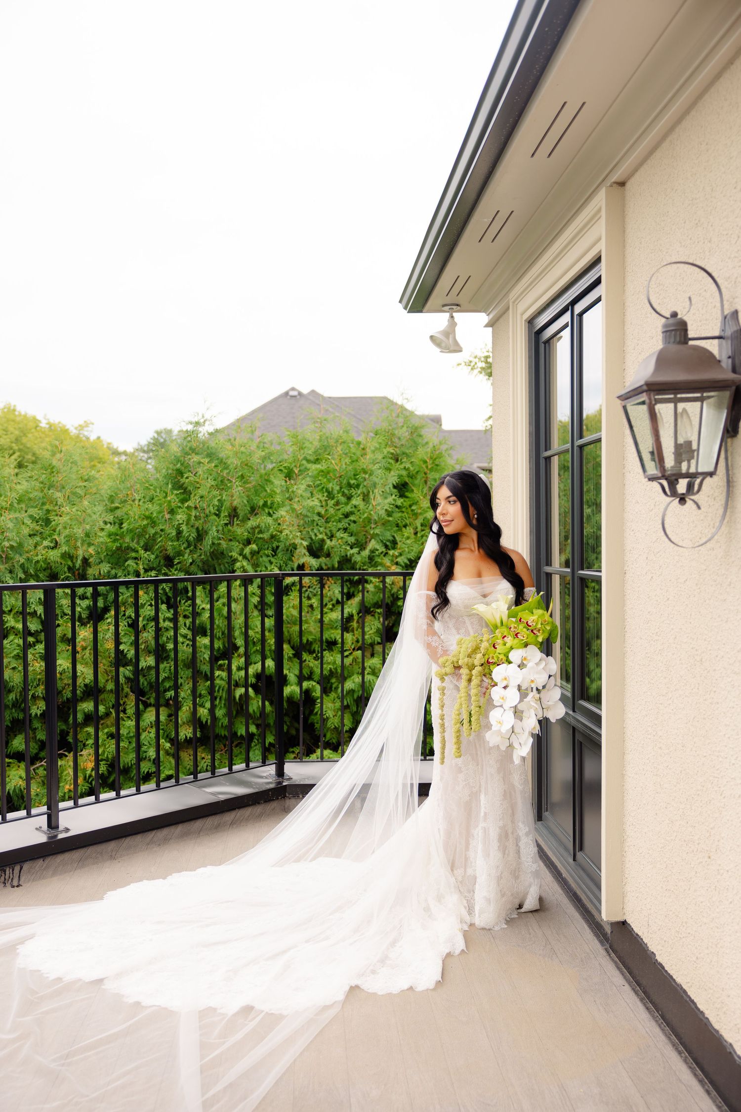 Bride in lace wedding gown with cascading veil stands on balcony holding elegant white and green floral bouquet.