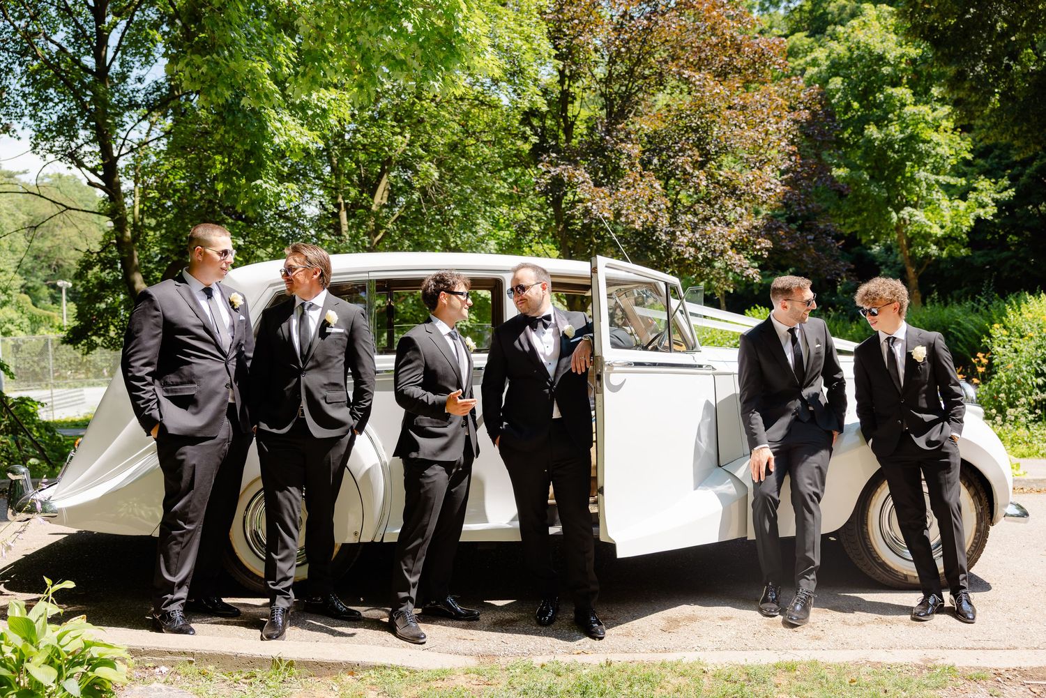 A wedding party in formal attire poses outdoors on a sunny day along a garden path in a candid sequence of shots.