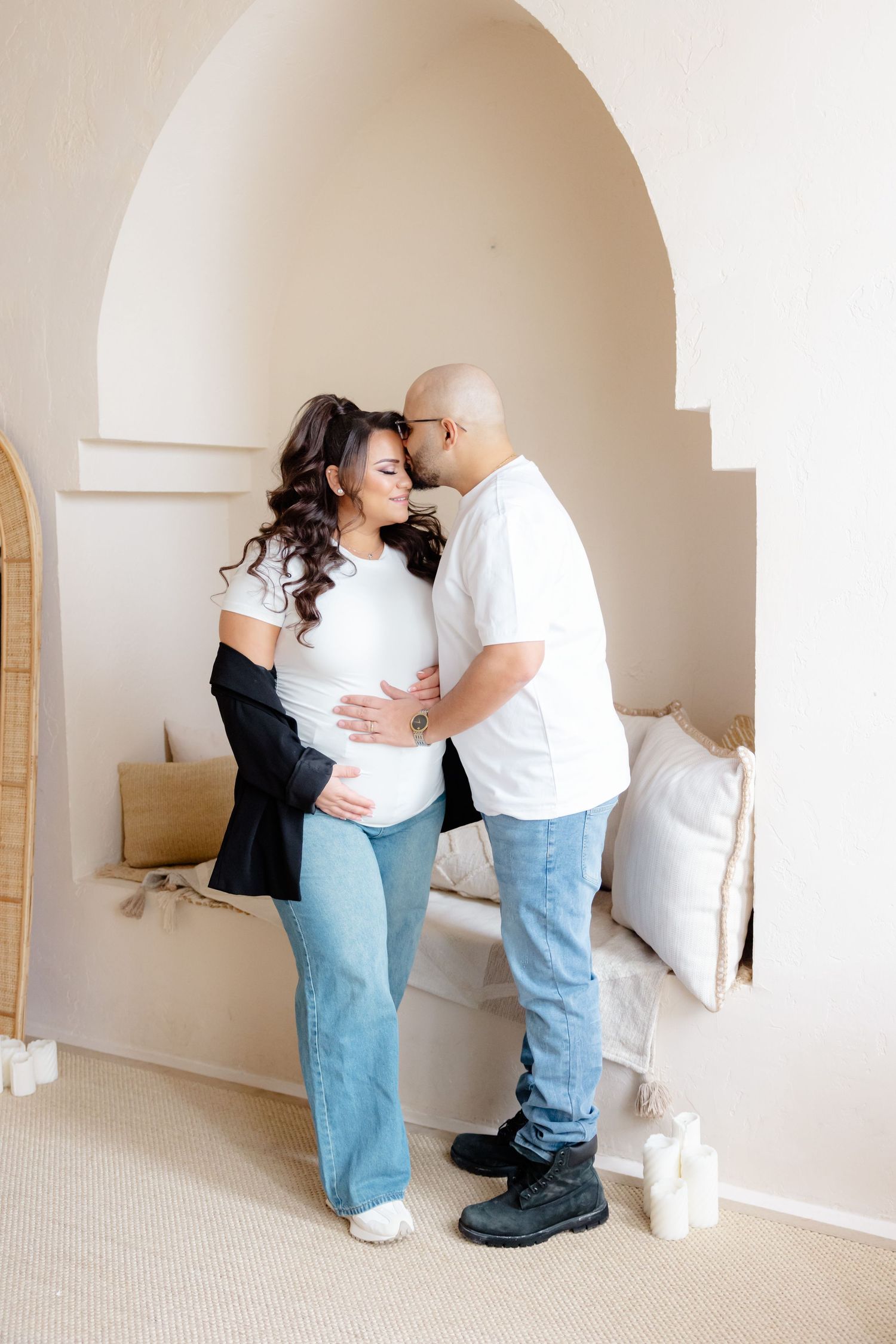 Expecting couple in white and denim share a tender moment in front of an arched doorway with light decor.