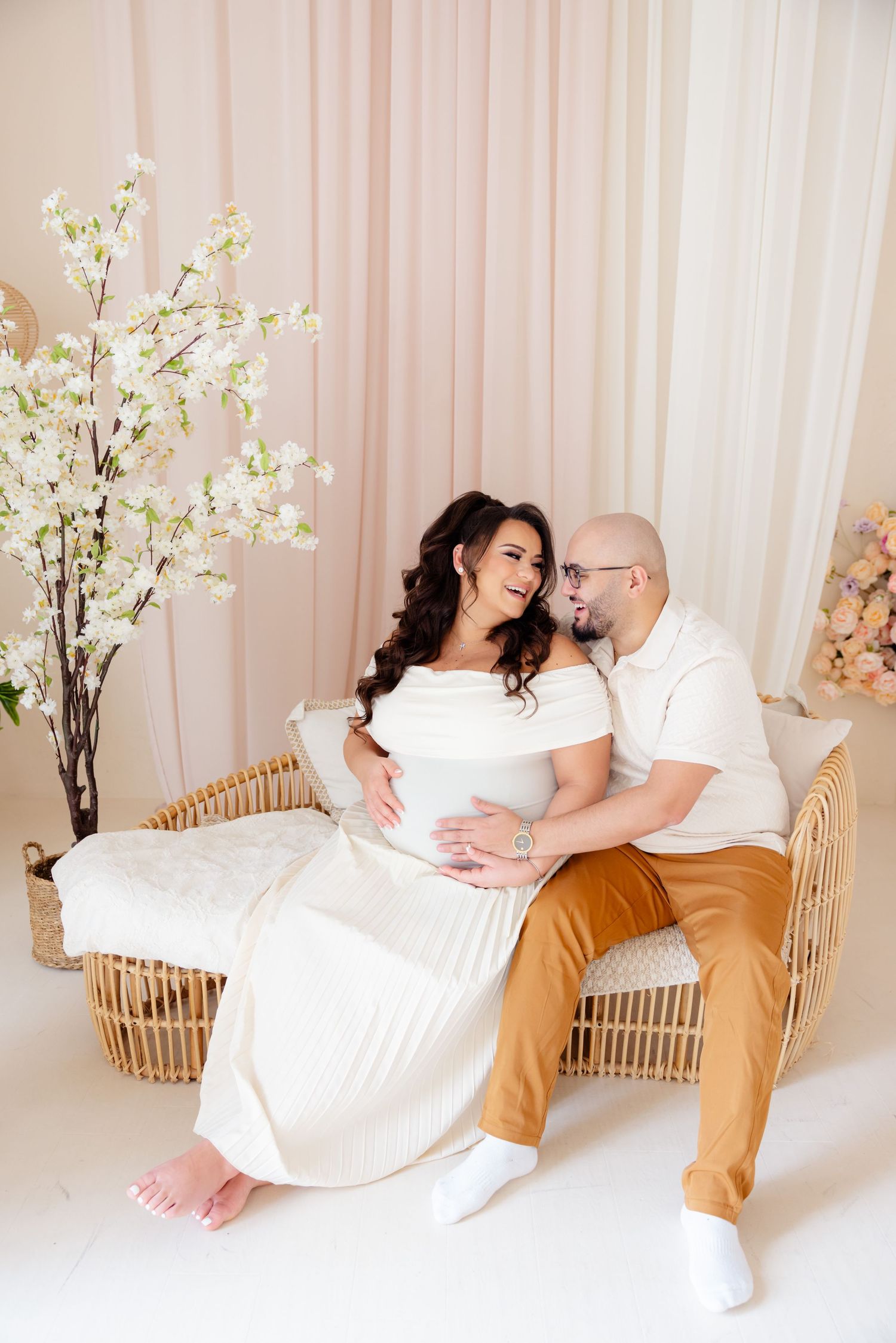 Expecting couple sits on wicker furniture with cherry blossoms in romantic pink studio setting.