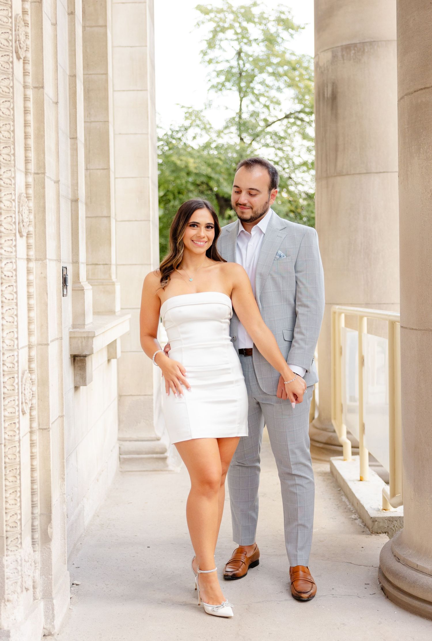 Stylish couple poses in formal attire between classic stone pillars.