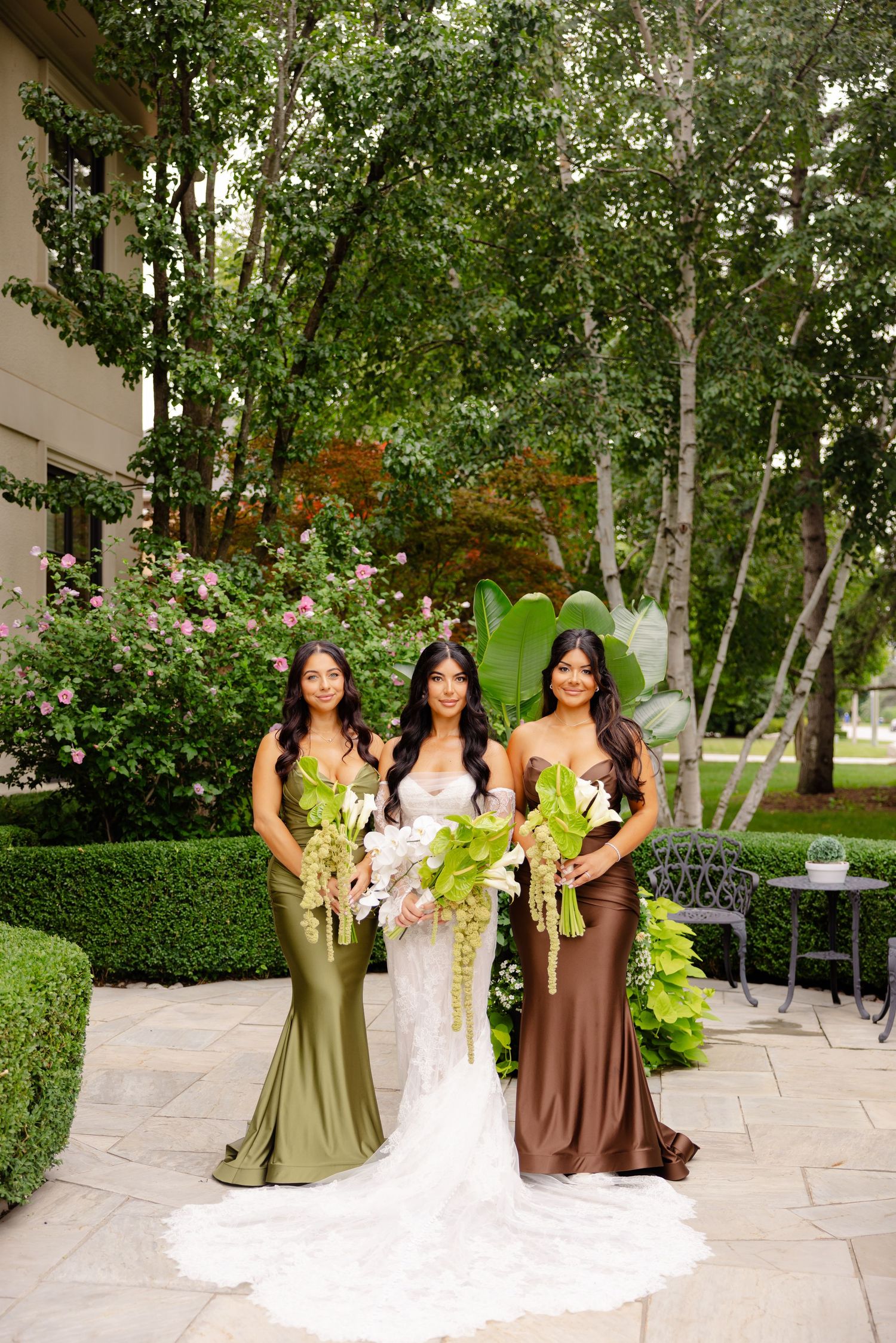Wedding party in earth-toned dresses poses on stone patio surrounded by lush greenery and trees.