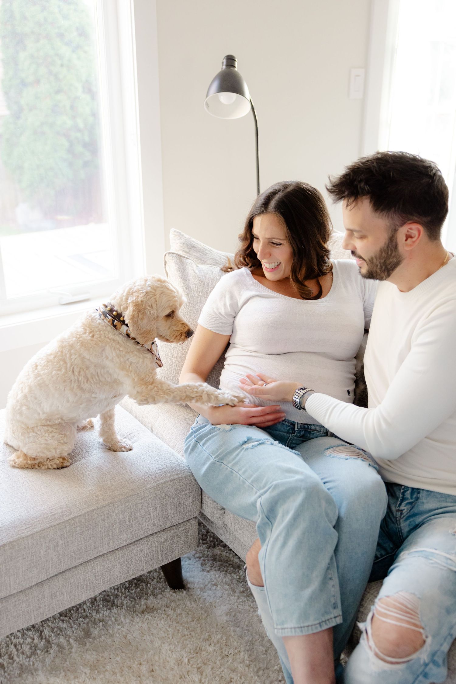 A couple sits on a couch with a small white dog, the woman is pregnant and they are smiling.