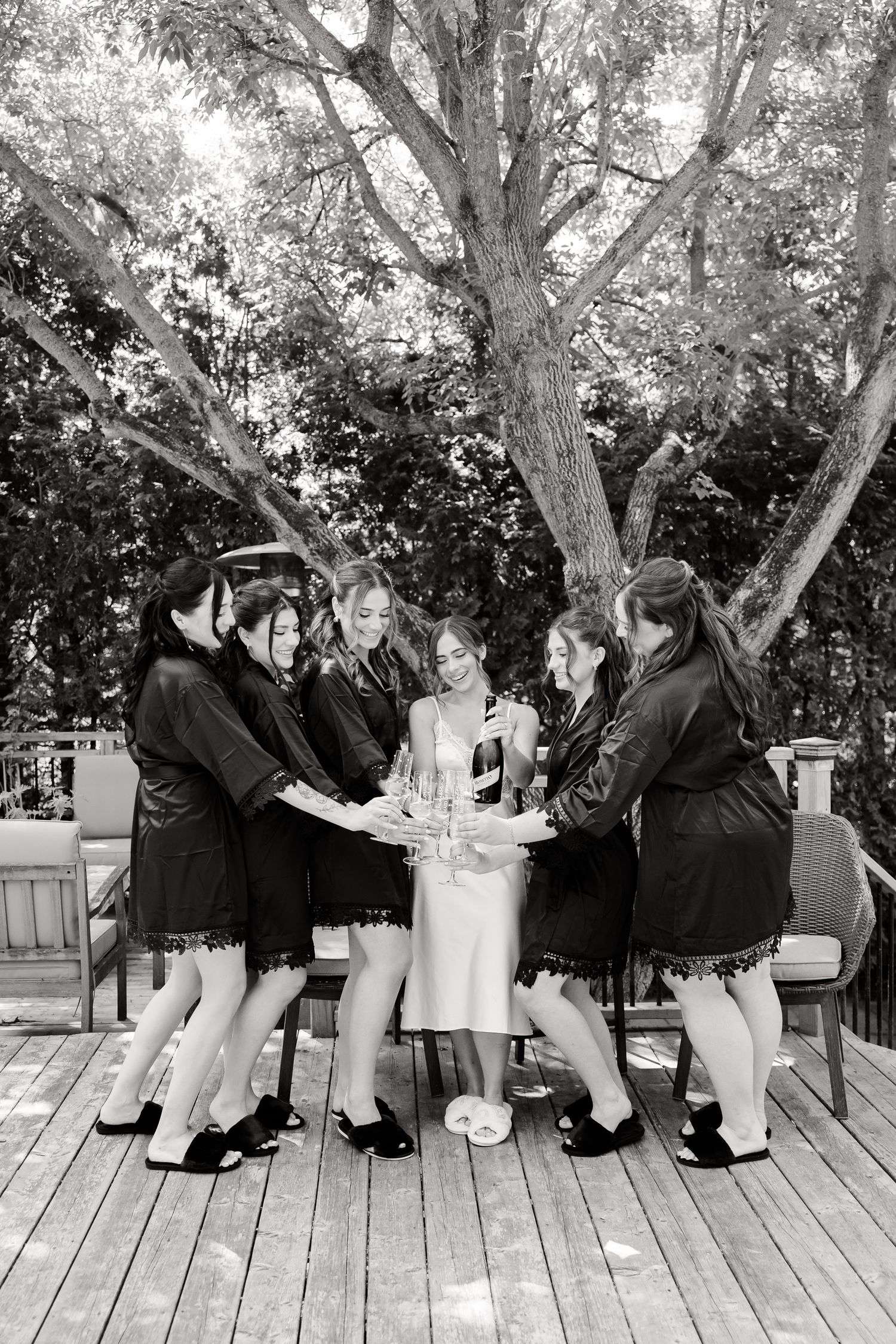 A black and white photo of a bridal party gathered in a circle on a wooden deck under trees during a celebration.