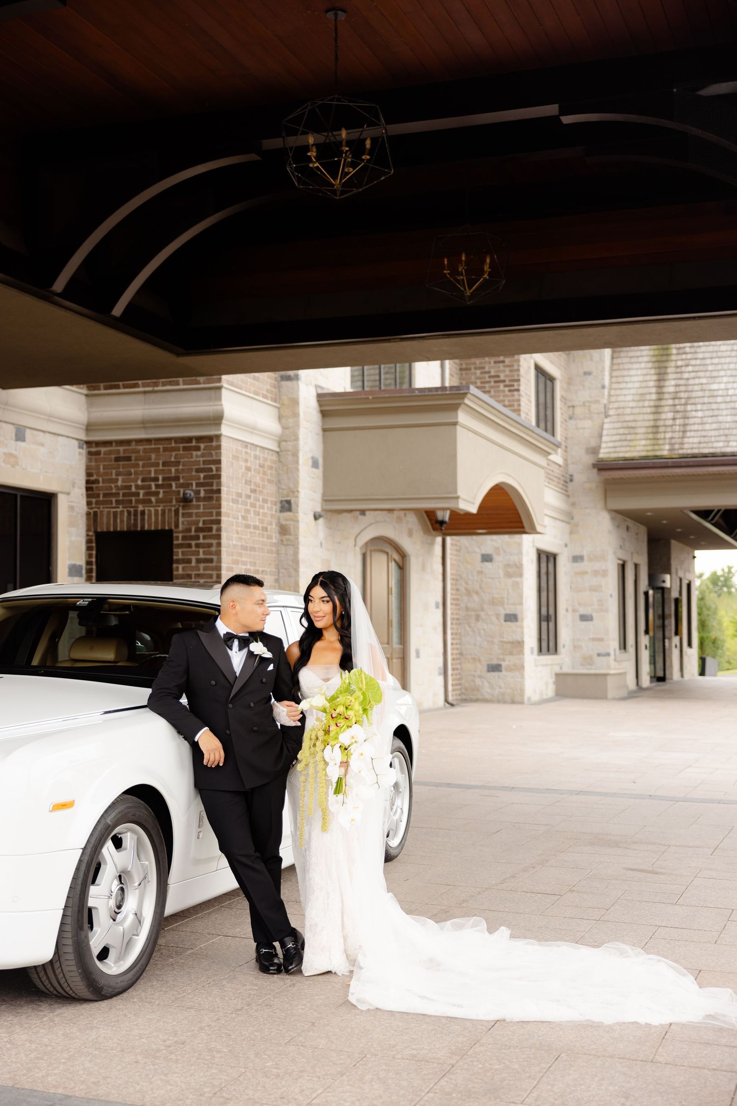 Elegant wedding couple poses near white luxury car under covered entrance of stone building.