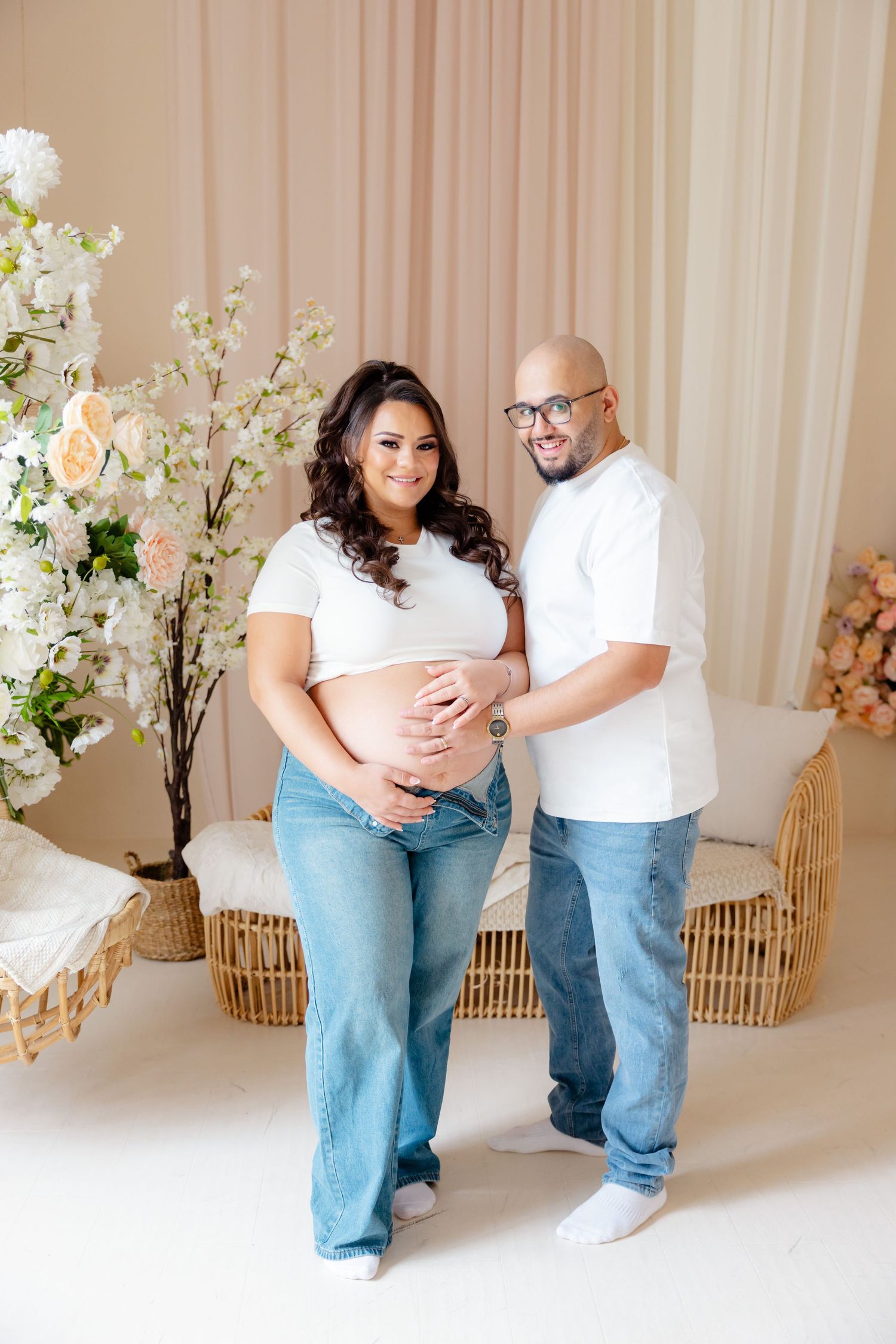 Expectant couple in white tops and jeans pose for maternity photo with floral decorations against beige backdrop.