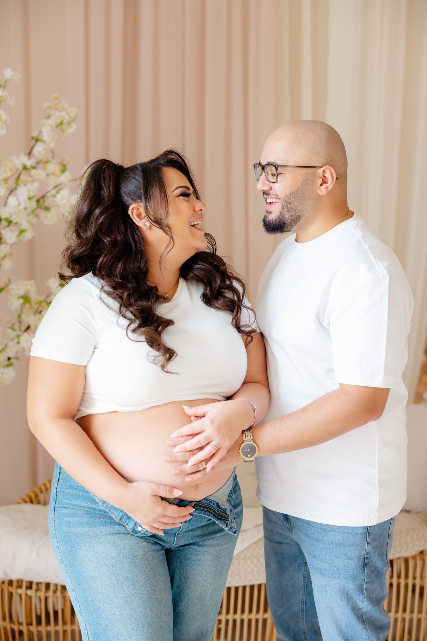 Happy couple wearing white shirts and jeans embrace while showing off her baby bump in a warm indoor setting.