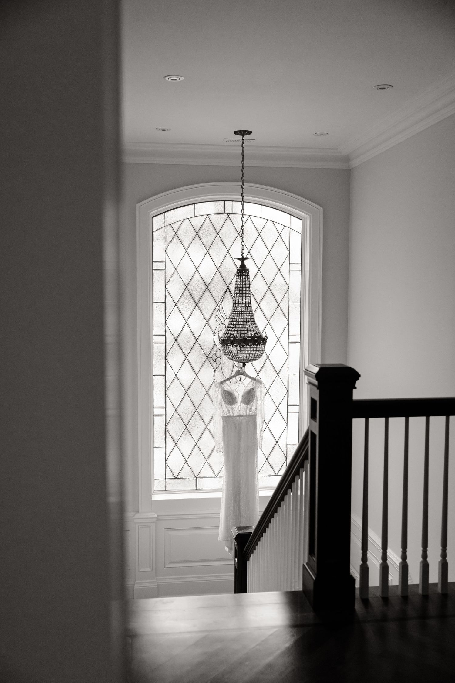 Black and white photo of wedding dress and chandelier against geometric window in stairwell.