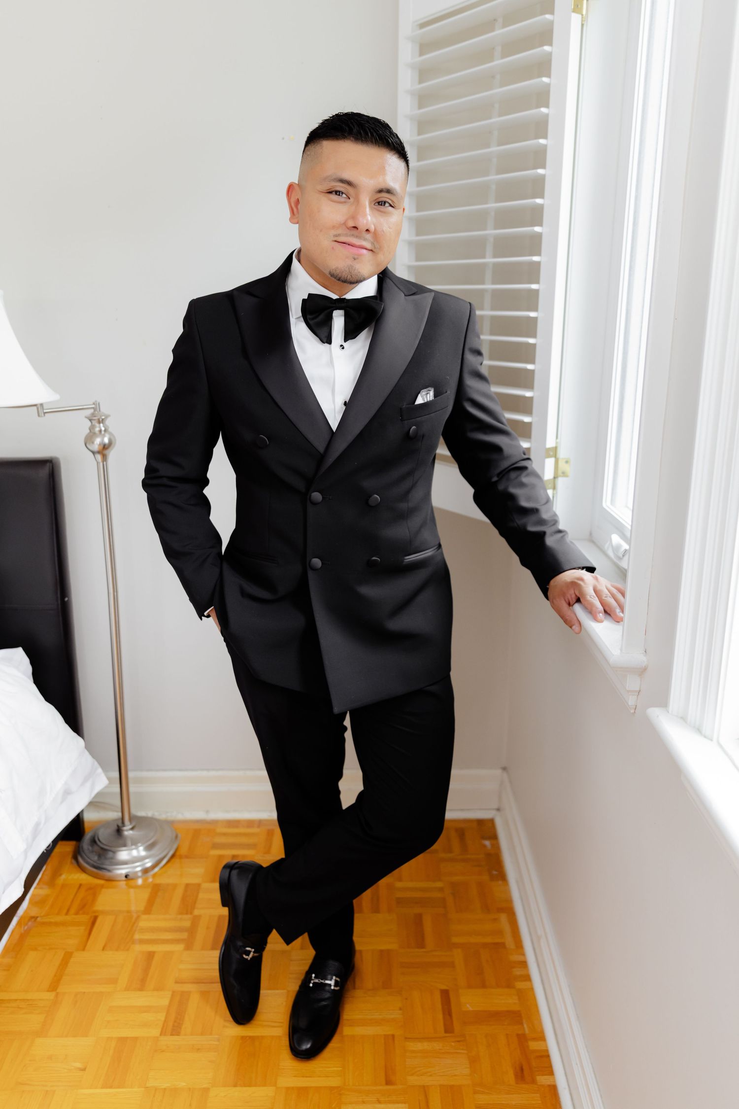 Person wearing a stylish black double-breasted tuxedo with bow tie stands elegantly against a white wall near a window.