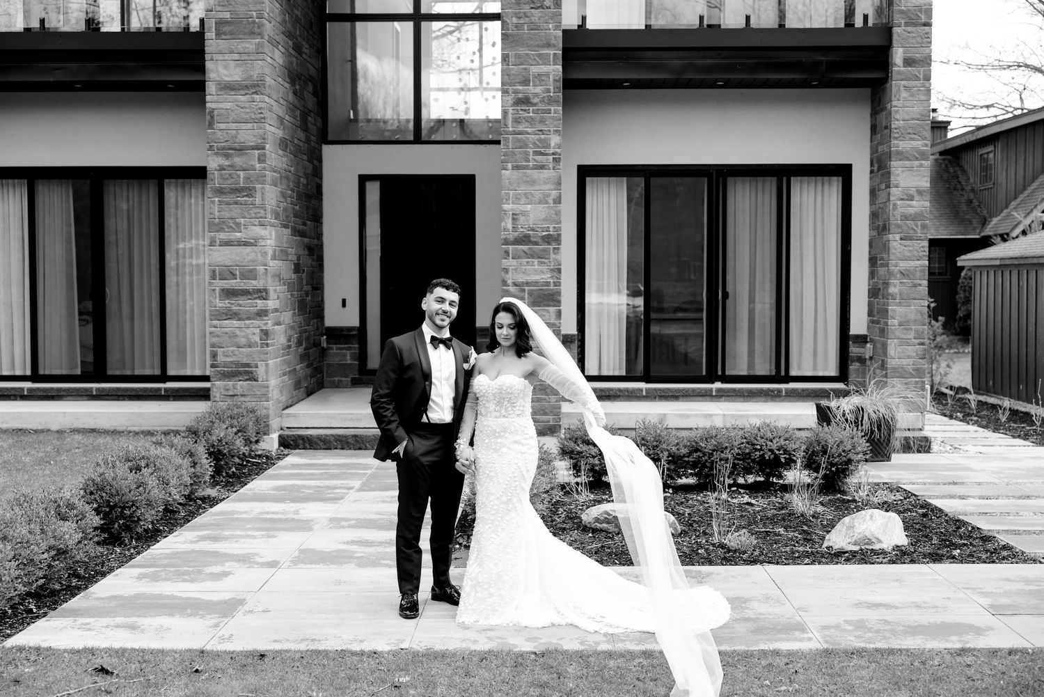 A newlywed couple walks in front of a modern stone building with their wedding attire gracefully flowing in black and white.