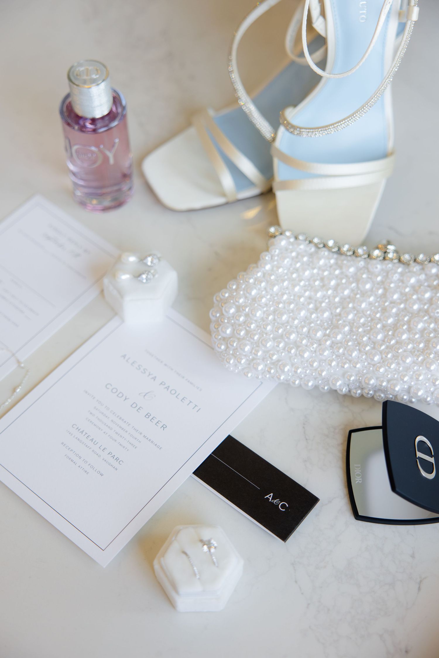 Wedding details flatlay featuring invitation, blue sandals and pearl clutch.