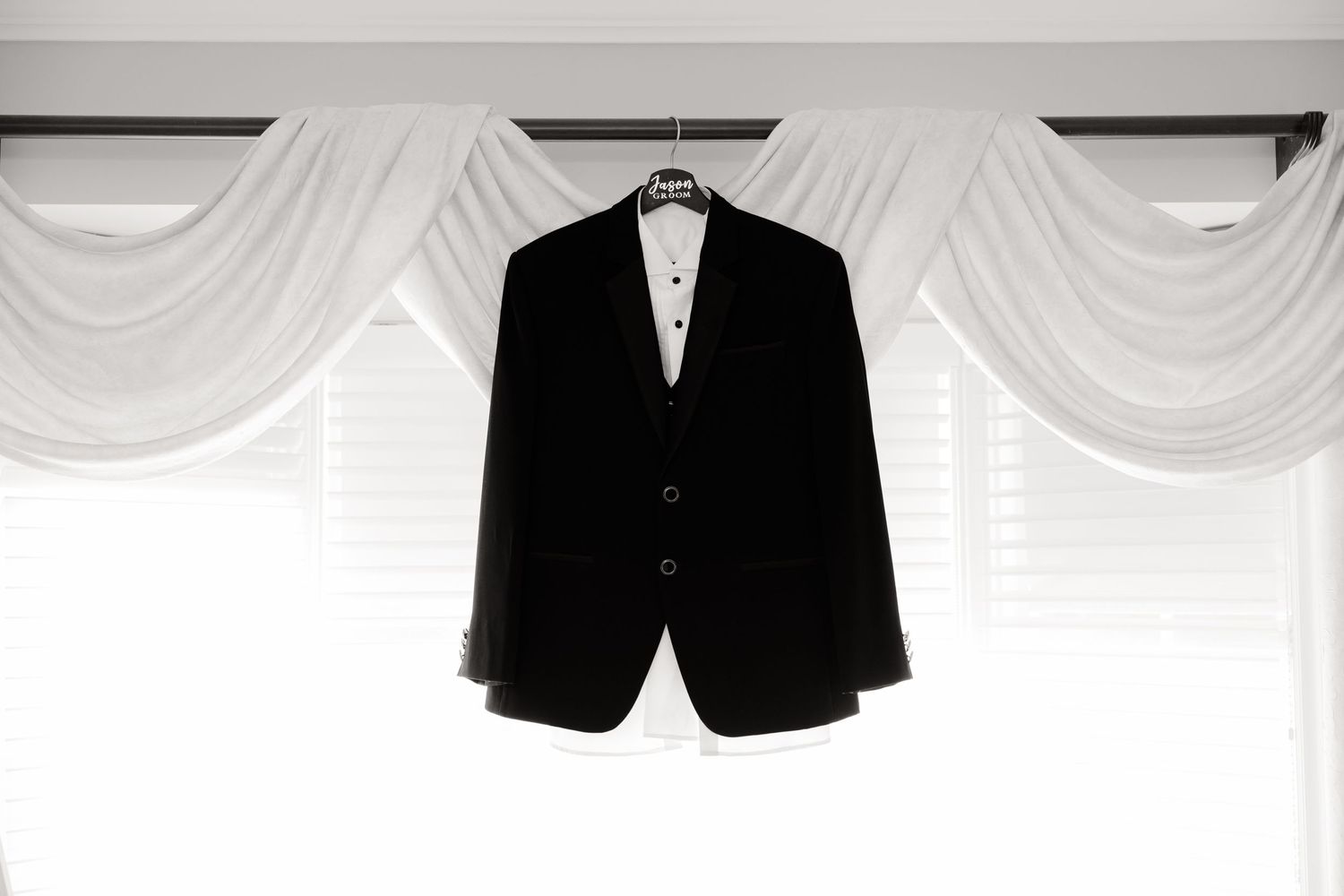 Black tuxedo jacket hanging against white draped curtains in formal setting.