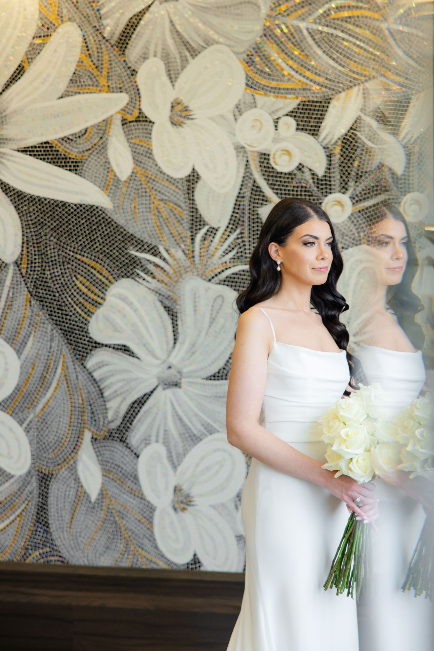 A bride in a sleek white gown holding a cream bouquet poses against a floral art wall with gold and gray details.