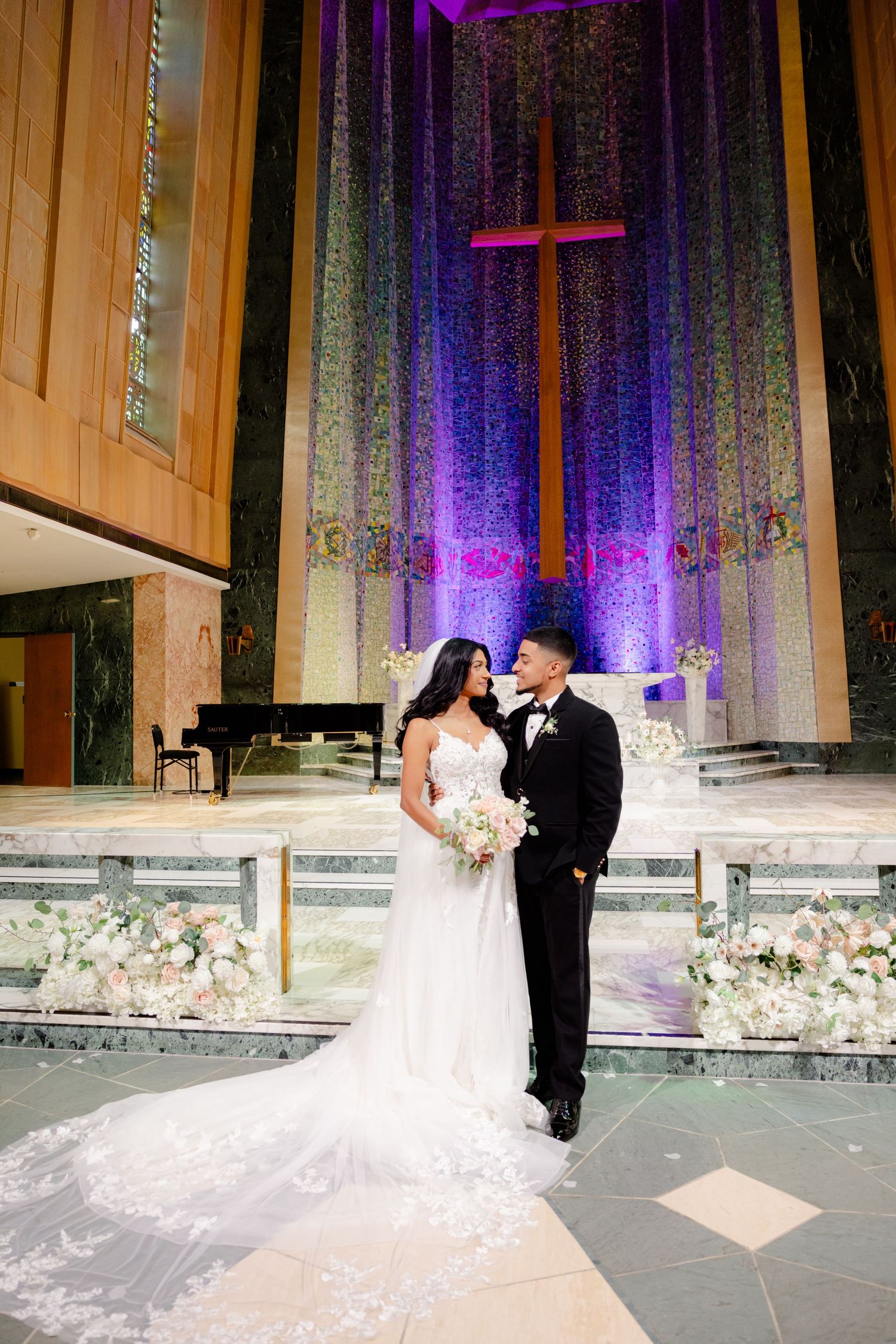 Intimate wedding portrait against purple-lit church altar with flowing white dress and florals.