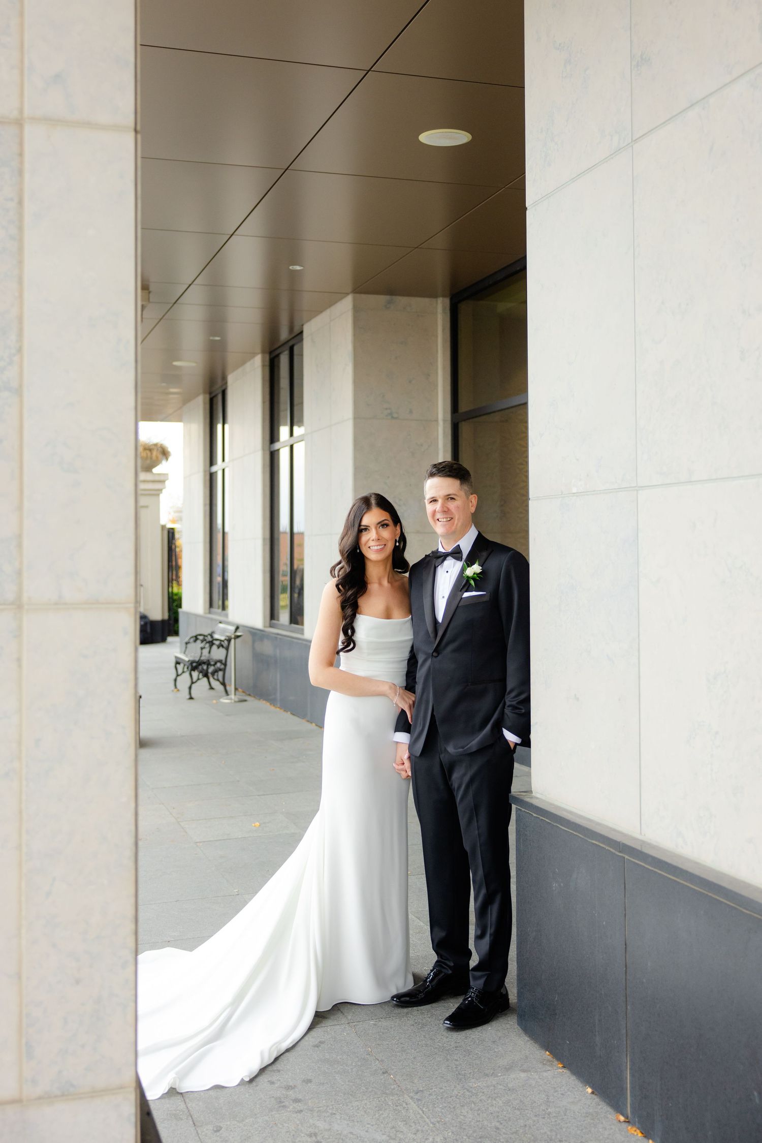 A romantic wedding portrait of a couple in formal attire standing together in an elegant covered walkway.