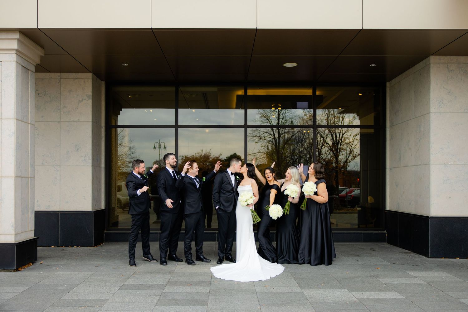 Wedding party gathered outside modern building entrance in formal black and white attire.