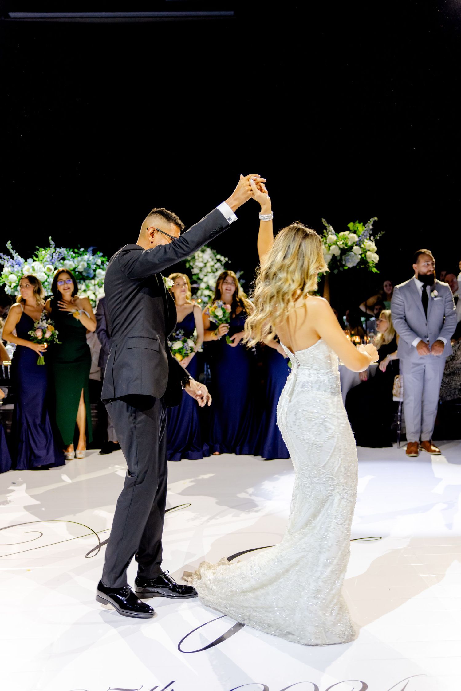 A wedding couple dancing on a white floor with bridesmaids in navy dresses in the background.