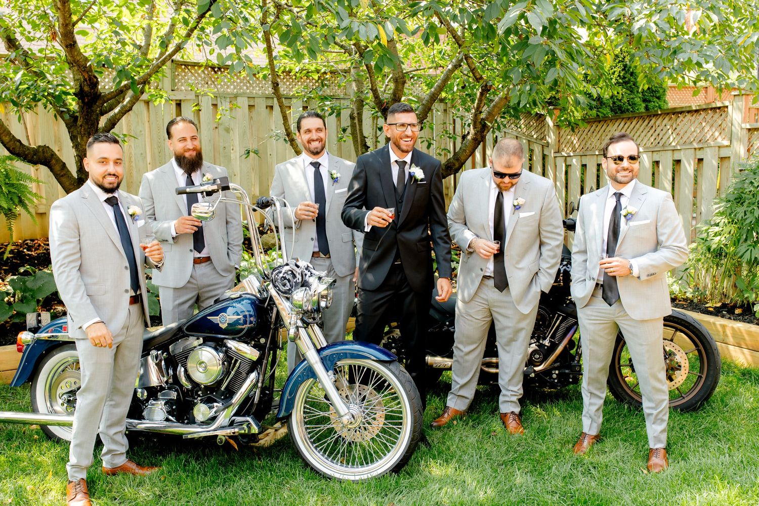 A group in light gray suits and dark ties pose with vintage motorcycles in a garden setting.