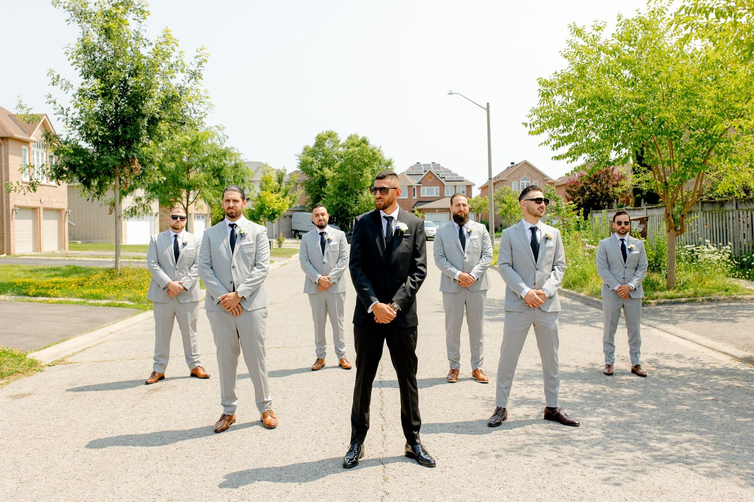 Wedding party in coordinated grey and black suits posing outdoors on suburban street.