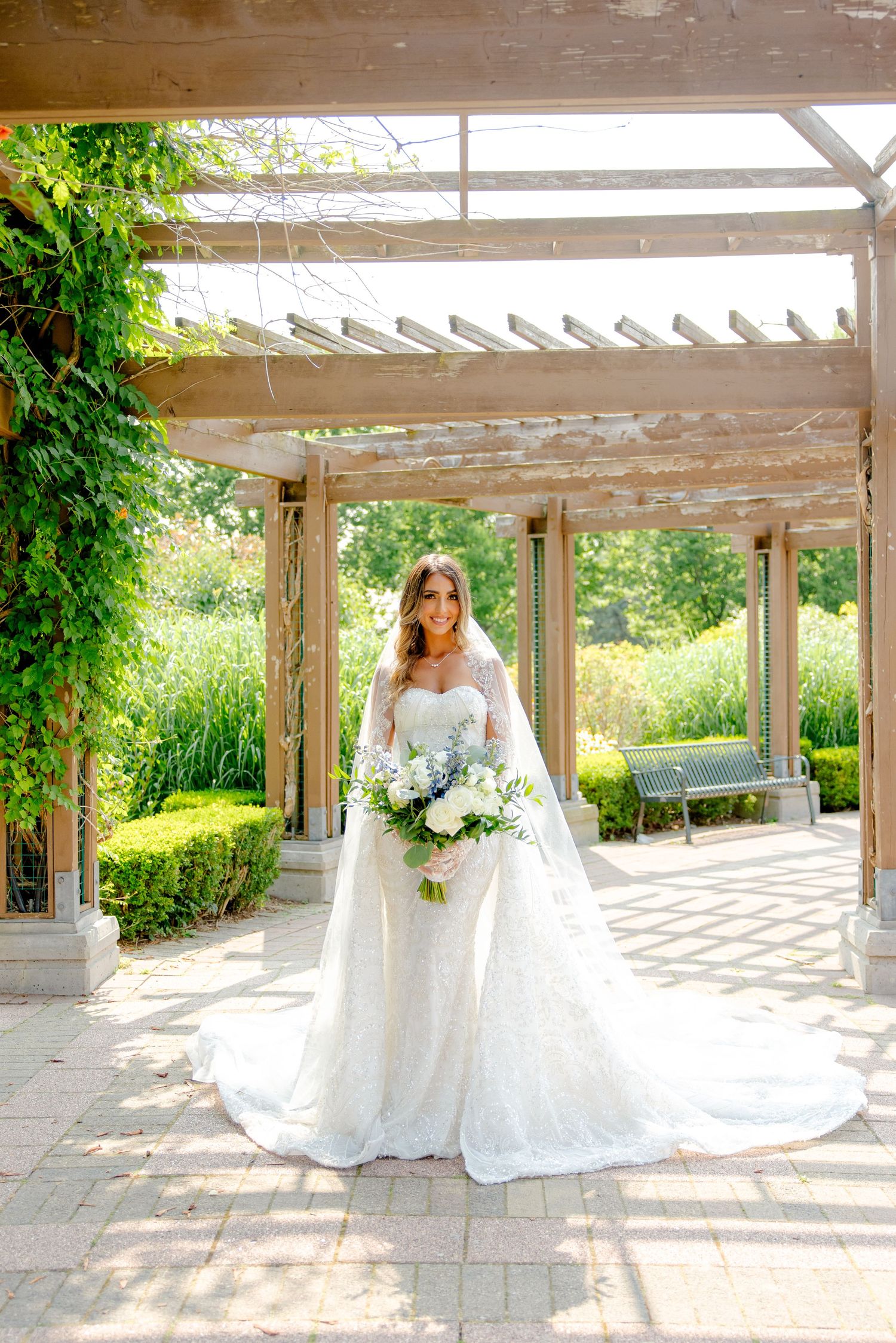 Bride in stunning white gown holds blue and white bouquet while posing under outdoor pergola.
