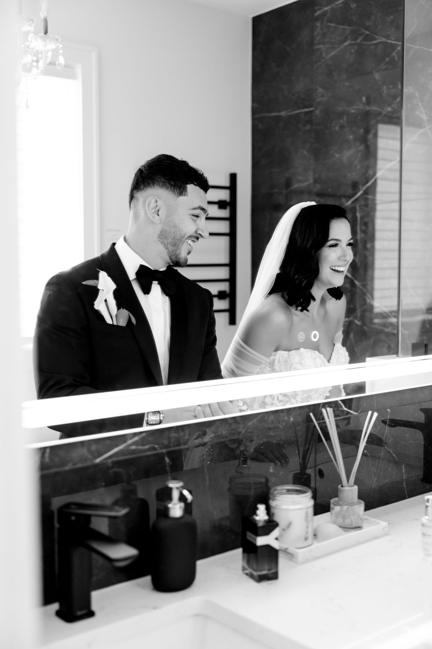 Black and white photo captures joyful wedding couple's reflection in a modern bathroom setting.