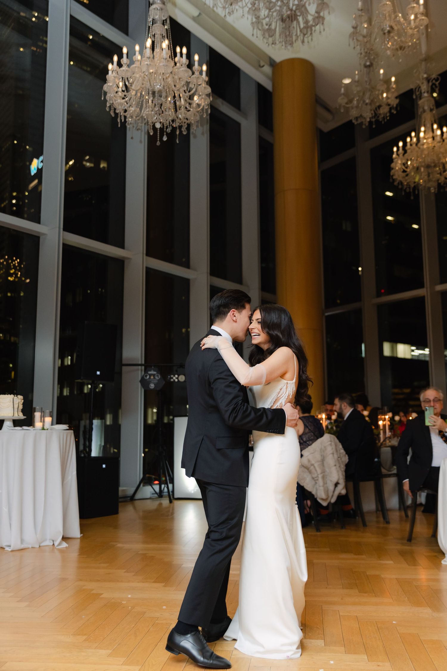 Romantic first dance beneath crystal chandeliers in modern venue with floor-to-ceiling windows.