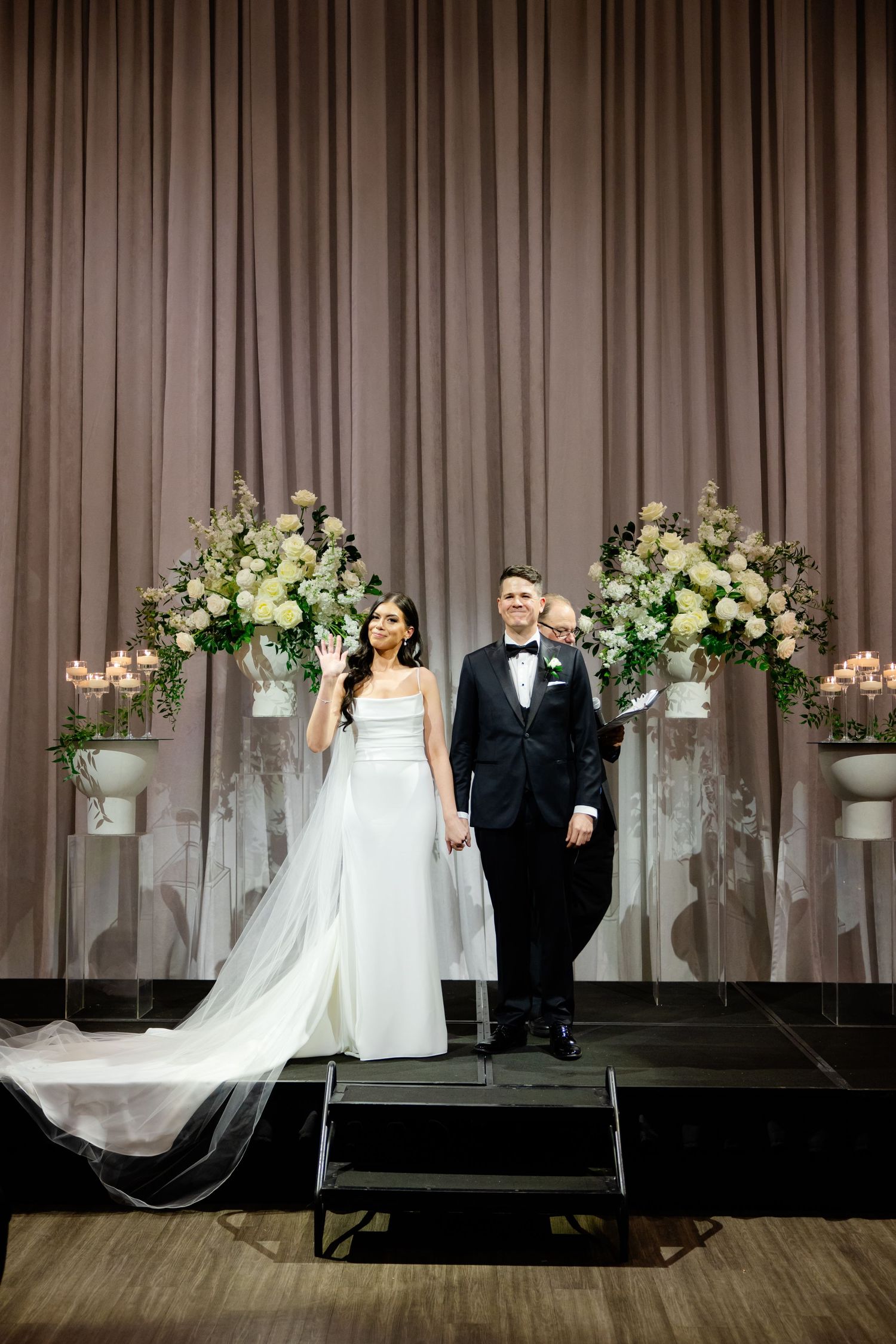 Elegant wedding portrait on stage with symmetrical white floral arrangements and flowing veil.